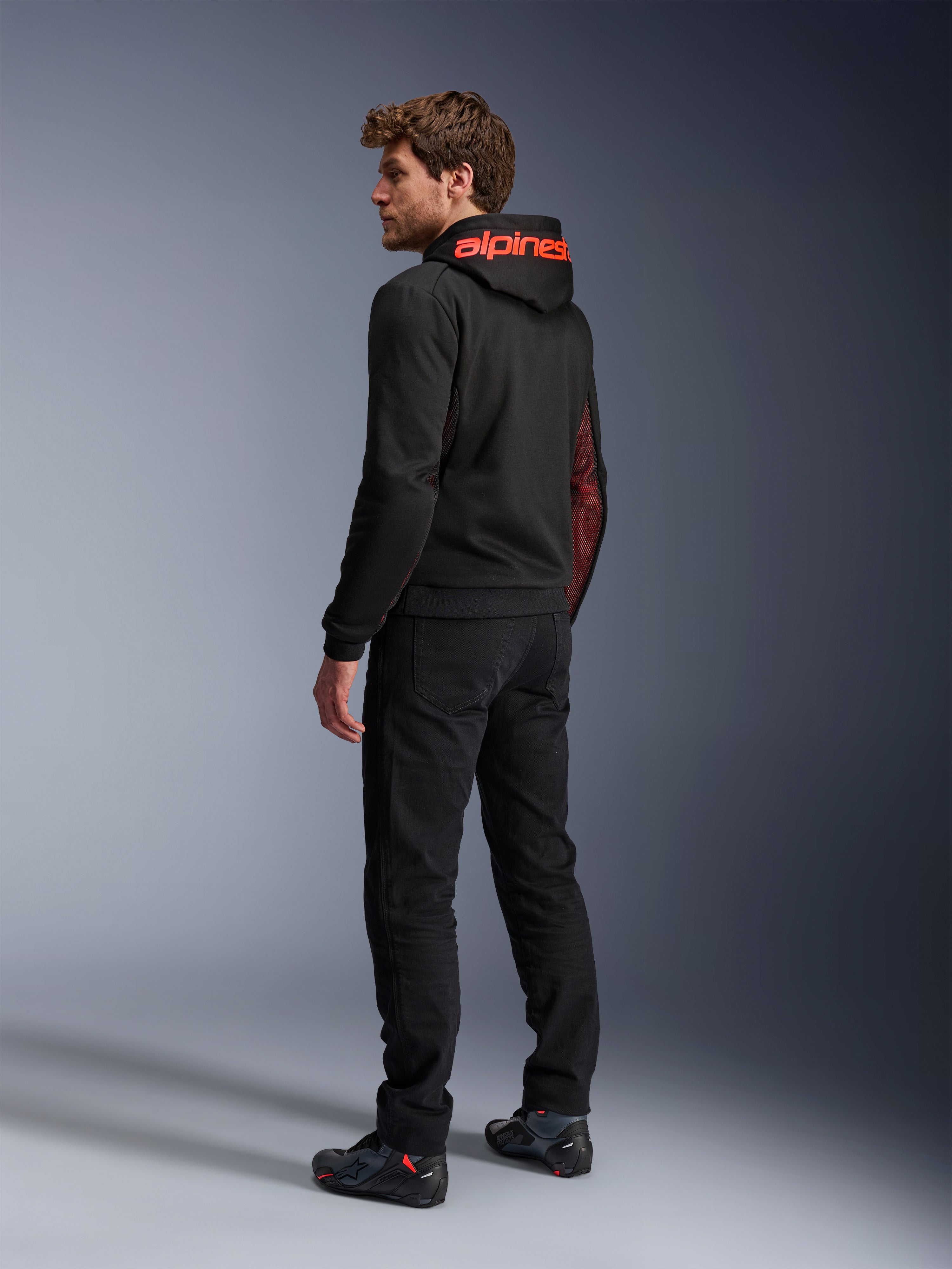 Alpinestars Chrome Air Sport Hoodie, urban motorcycle hoodie, Black and Red Fluo, rear view showing red Alpinestars branding on the hood and mesh side panels, worn with black riding pants and technical footwear