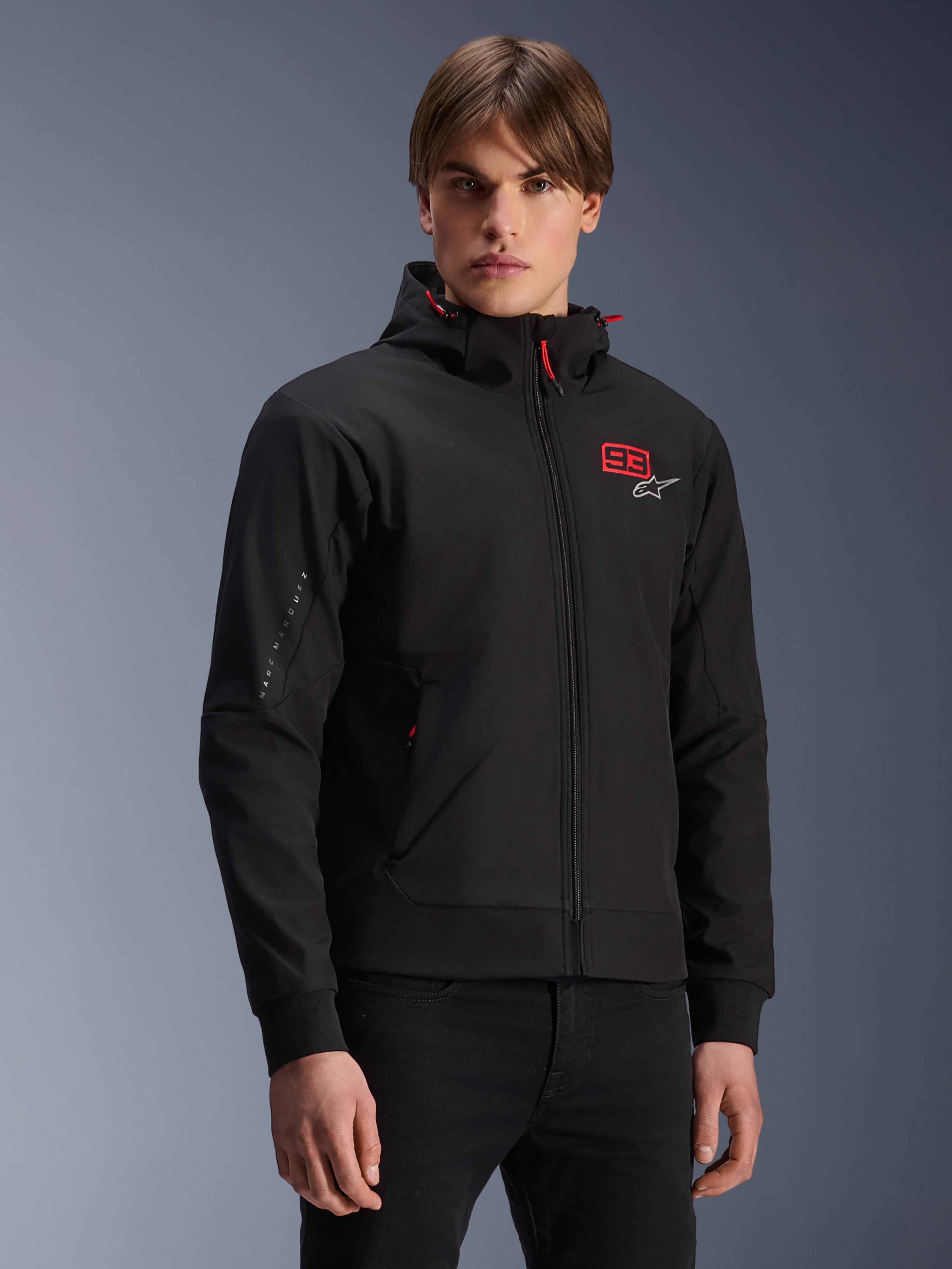 MM93 Radium Tech Hoodie, schwarze urbane Motorrad-Kapuzenjacke mit Reißverschluss, rote 93 und weißes Alpinestars-Logo auf der Brust, von einem Model mit schwarzer Hose getragen, abnehmbare Kapuze mit roten Kordelzügen und Branding am Ärmel, lässiges vom Motorsport inspiriertes Design