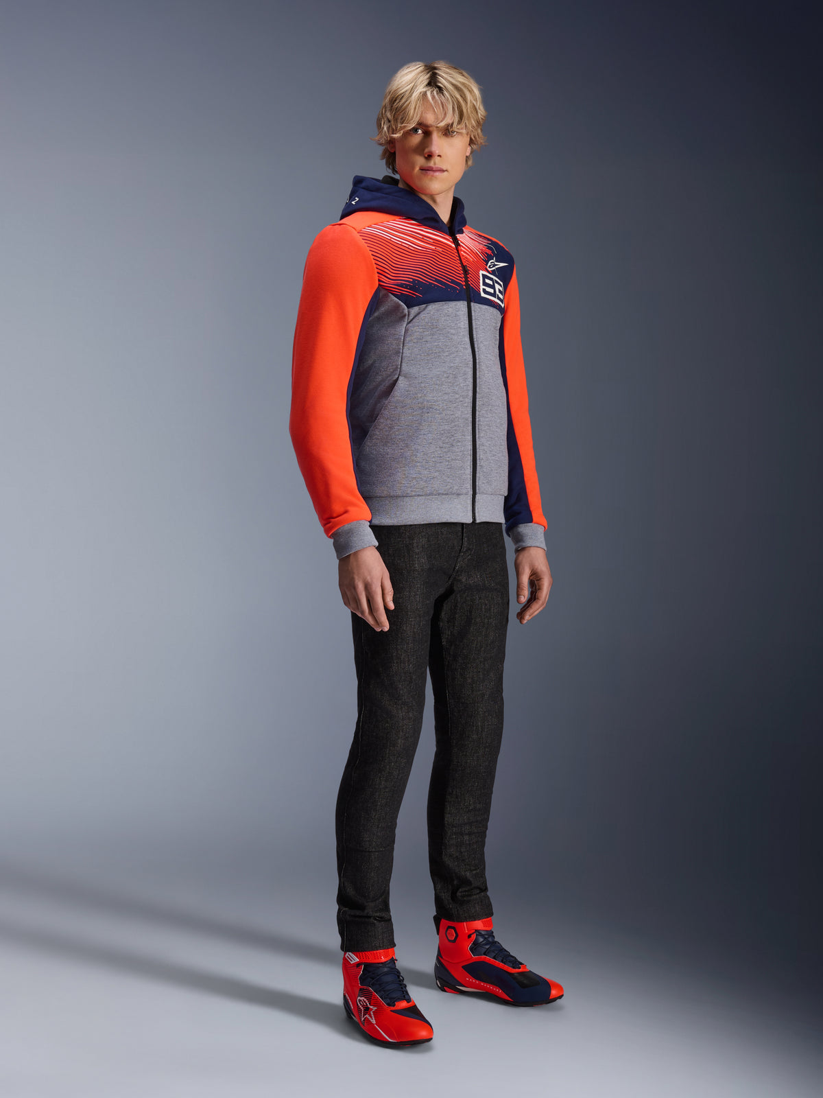 Alpinestars MM93 Chrome V2 Sport Hoodie, urbaner Motorrad-Hoodie, grau melierter Hauptteil mit marineblauer Brust und leuchtend neonroten Ärmeln, 93-Logo auf der Brust, getragen von einem Model mit schwarzer Slim-Fit-Hose und passenden rot-blauen High-Top-Fahrschuhen, moderne athletische Passform für den Einsatz in der Stadt