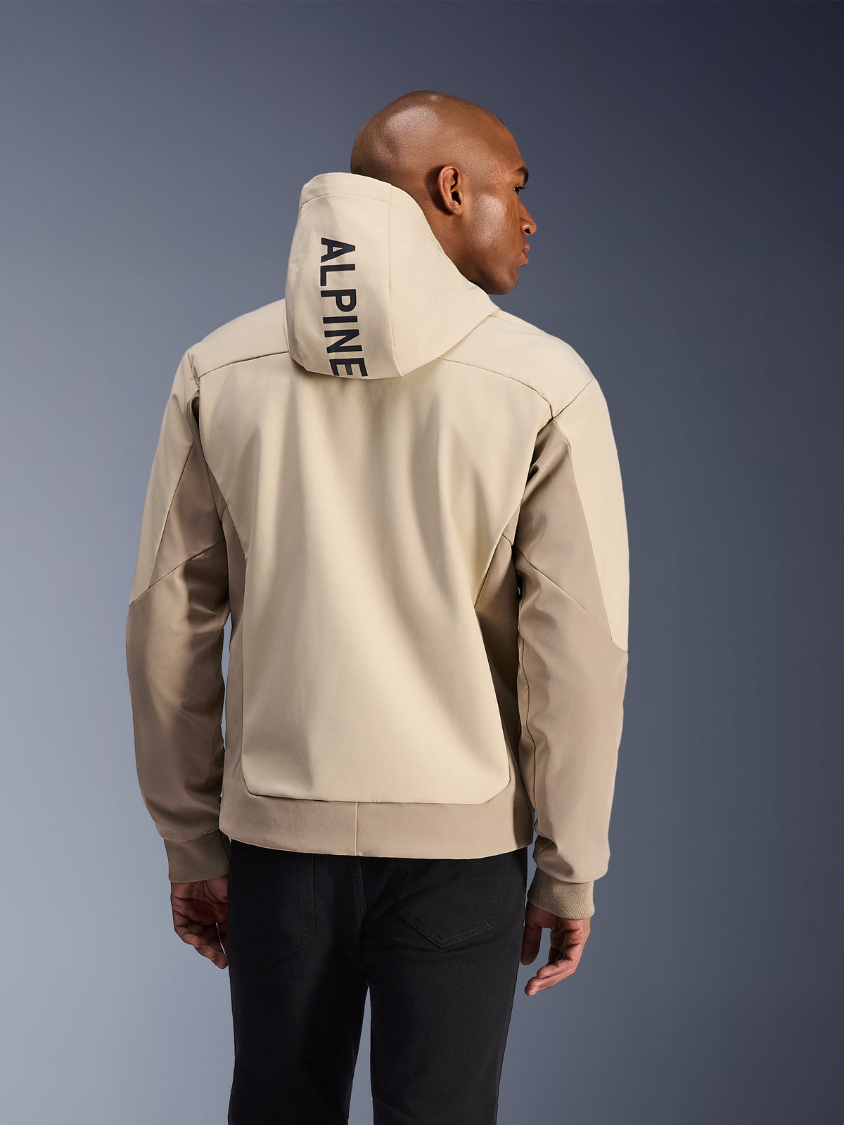 Alpinestars Radium Tech Hoodie, Rückansicht eines Mannes im technischen Hoodie in Mastic Aluminum, vertikaler ALPINE-Schriftzug auf der Kapuze, hellbeige und sandfarbene Panele, gerippte Bündchen, urbane Motorradjacke