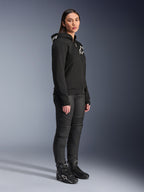 Woman wearing Alpinestars Stella Chrome V2 Sport Hoodie in black, featuring white Alpinestars branding, paired with black motorcycle riding pants and boots, urban riding gear for women with a casual athletic fit