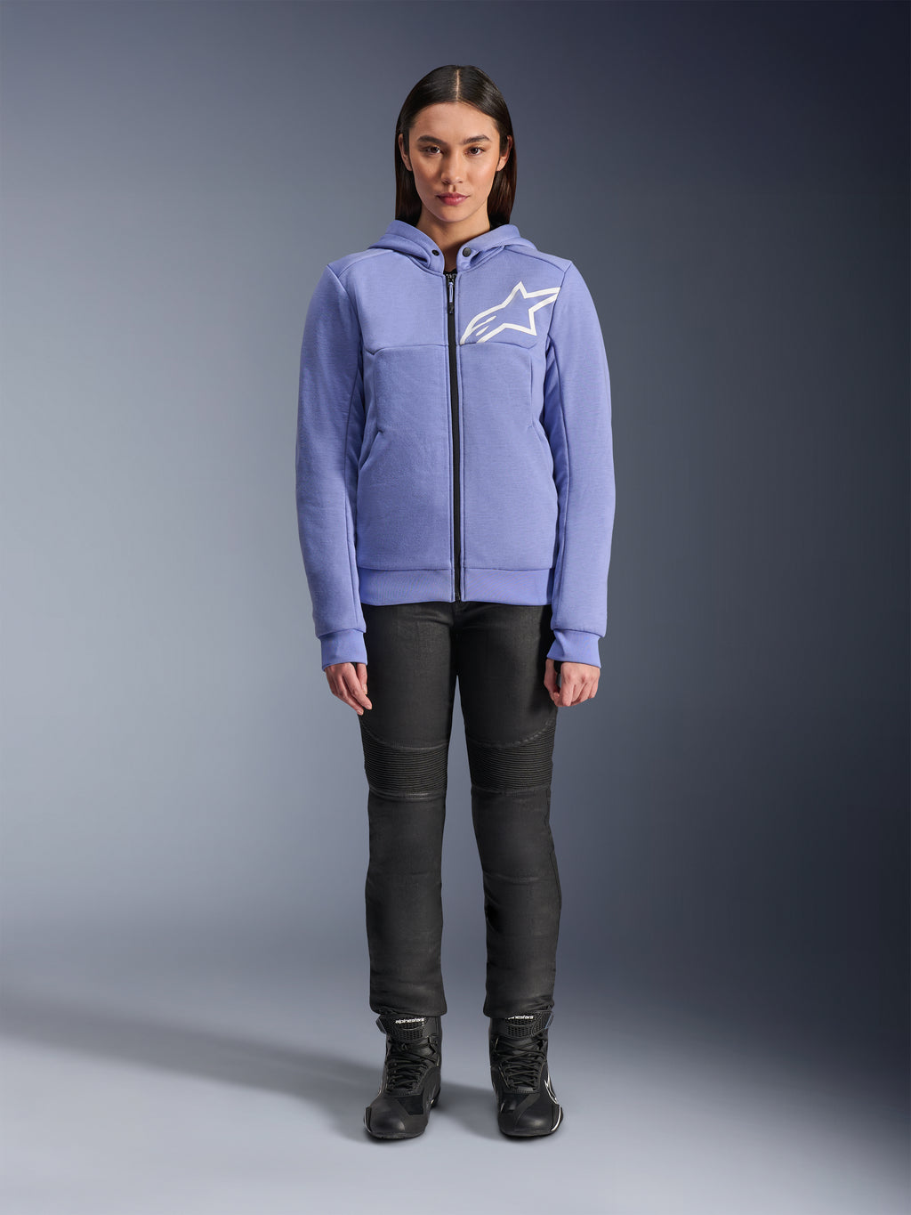 Alpinestars Stella Chrome V2 Sport Damen-Hoodie, Blau-Violett mit weißem Astars-Logo auf der Brust, technisches Fleece mit durchgehendem Reißverschluss und Kapuze, getragen mit schwarzer Motorradhose und Motorradstiefeln, lässige urbane Motorradbekleidung für Damen