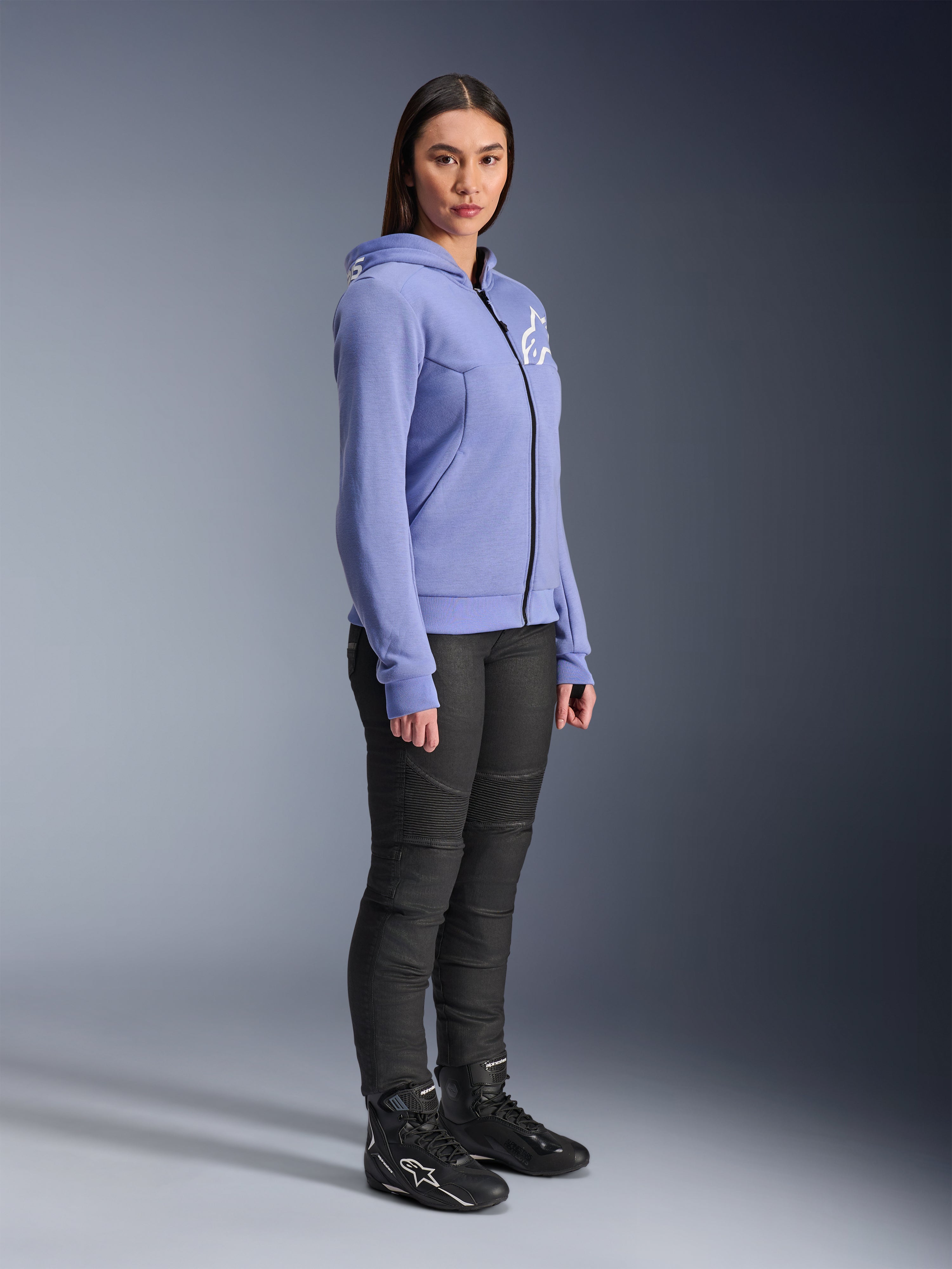 Alpinestars Stella Chrome V2 Sport Damen-Hoodie, Motorrad-Hoodie in Blau-Violett mit weißem Alpinestars-Logo auf der Brust, getragen von einem Modell mit schwarzer Motorradhose und schwarzen Motorradstiefeln, urbane Motorradbekleidung für Damen
