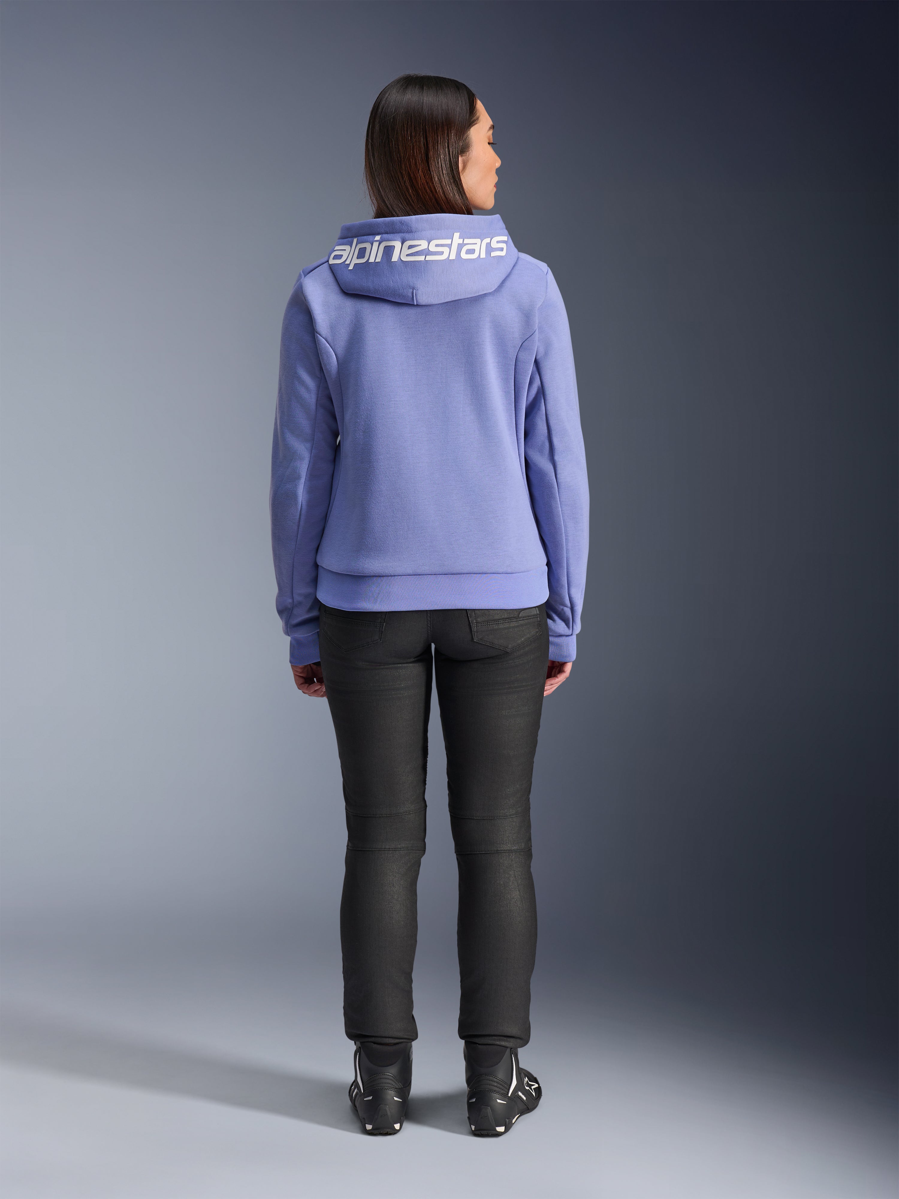 Alpinestars Stella Chrome V2 Sport Hoodie, Blau-Violett, Rückansicht einer Frau, die den Hoodie mit weißem Alpinestars-Logo auf der Kapuze trägt, kombiniert mit schwarzer Hose und Motorradstiefeln, lässige urbane Motorradbekleidung