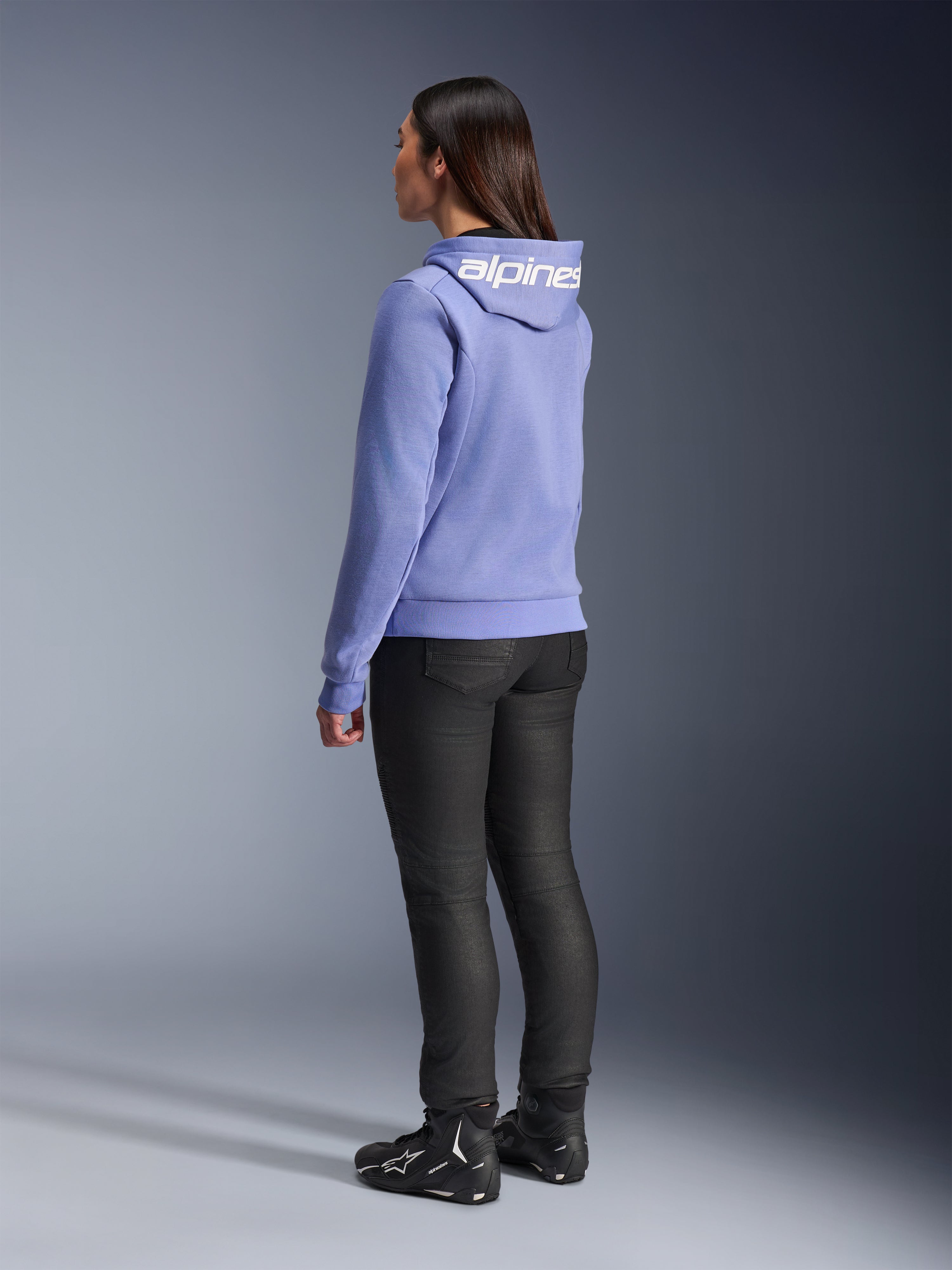 Alpinestars Stella Chrome V2 Sport Damen-Hoodie, Blau-Violett, Rückansicht mit weißem Branding auf der Kapuze, getragen mit schwarzer Motorradhose und schwarzen Motorradstiefeln, lässige urbane Motorradausrüstung