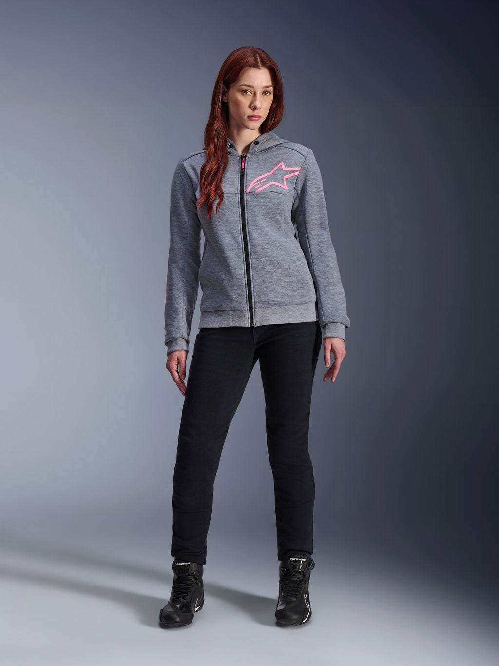 Alpinestars Stella Chrome V2 Sport Hoodie, Gray Heather Pink Fluo, getragen von einer Frau mit schwarzer Hose und Stiefeln, mit einem pinkfarbenen Alpinestars Stern-Logo auf der Brust, Urban-Motorradbekleidung mit durchgehendem Reißverschluss, leichtes Tech-Fleece mit lässiger, sportlicher Passform.