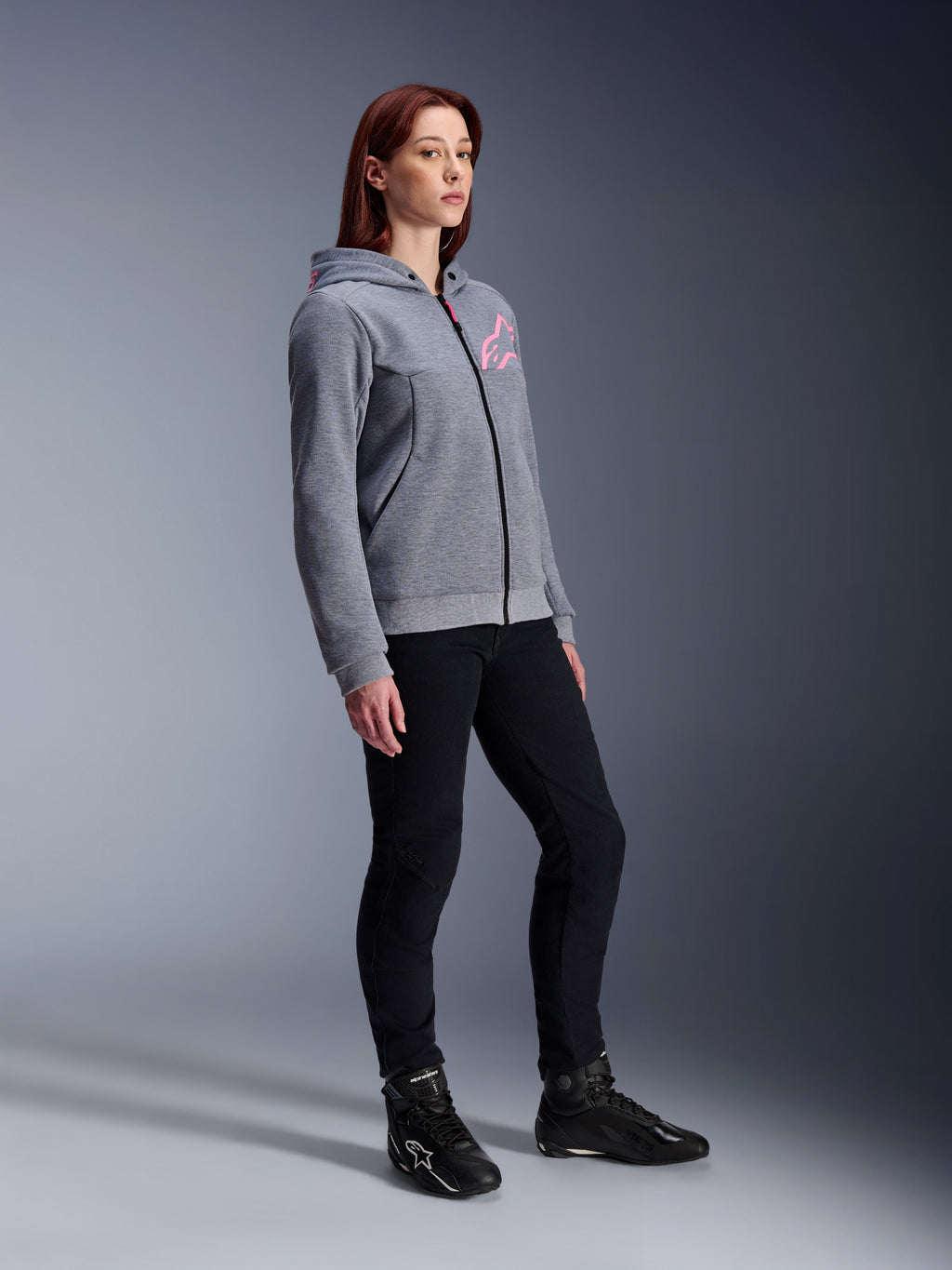 Alpinestars Stella Chrome V2 Sport Hoodie, Motorrad-Kapuzenjacke für Damen, Grau-Meliert und Neon-Pink, getragen mit schwarzer Motorradhose und schwarzen Stiefeln, lässige urbane Motorradausrüstung