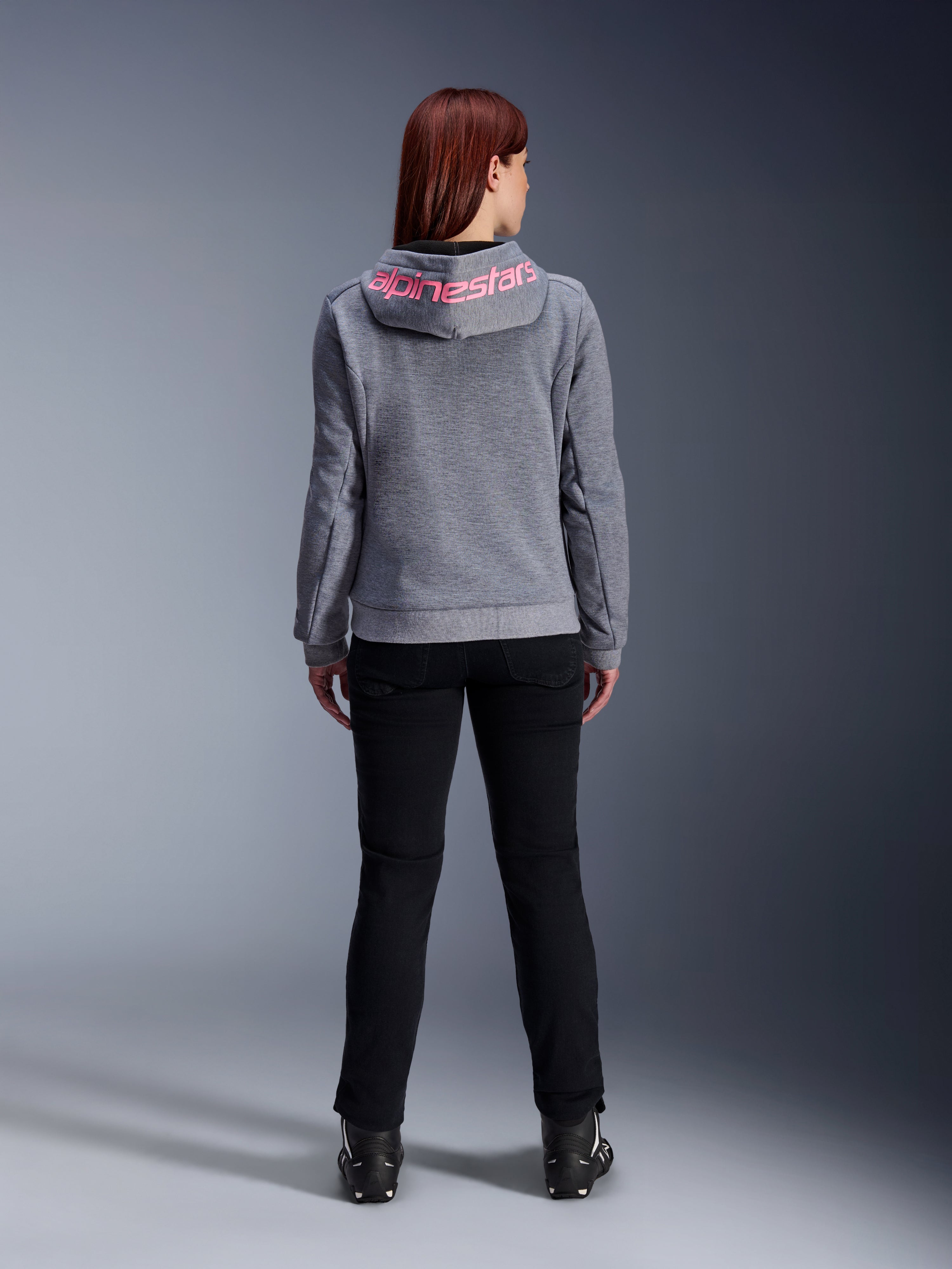 Alpinestars Stella Chrome V2 Sport Hoodie, Motorrad-Kapuzenjacke für Damen, Grau Meliert und Pink Fluo, Rückansicht mit pinkem Alpinestars-Logo auf der Kapuze, getragen mit schwarzer Motorradhose und Stiefeln, lässige Motorradbekleidung für Damen