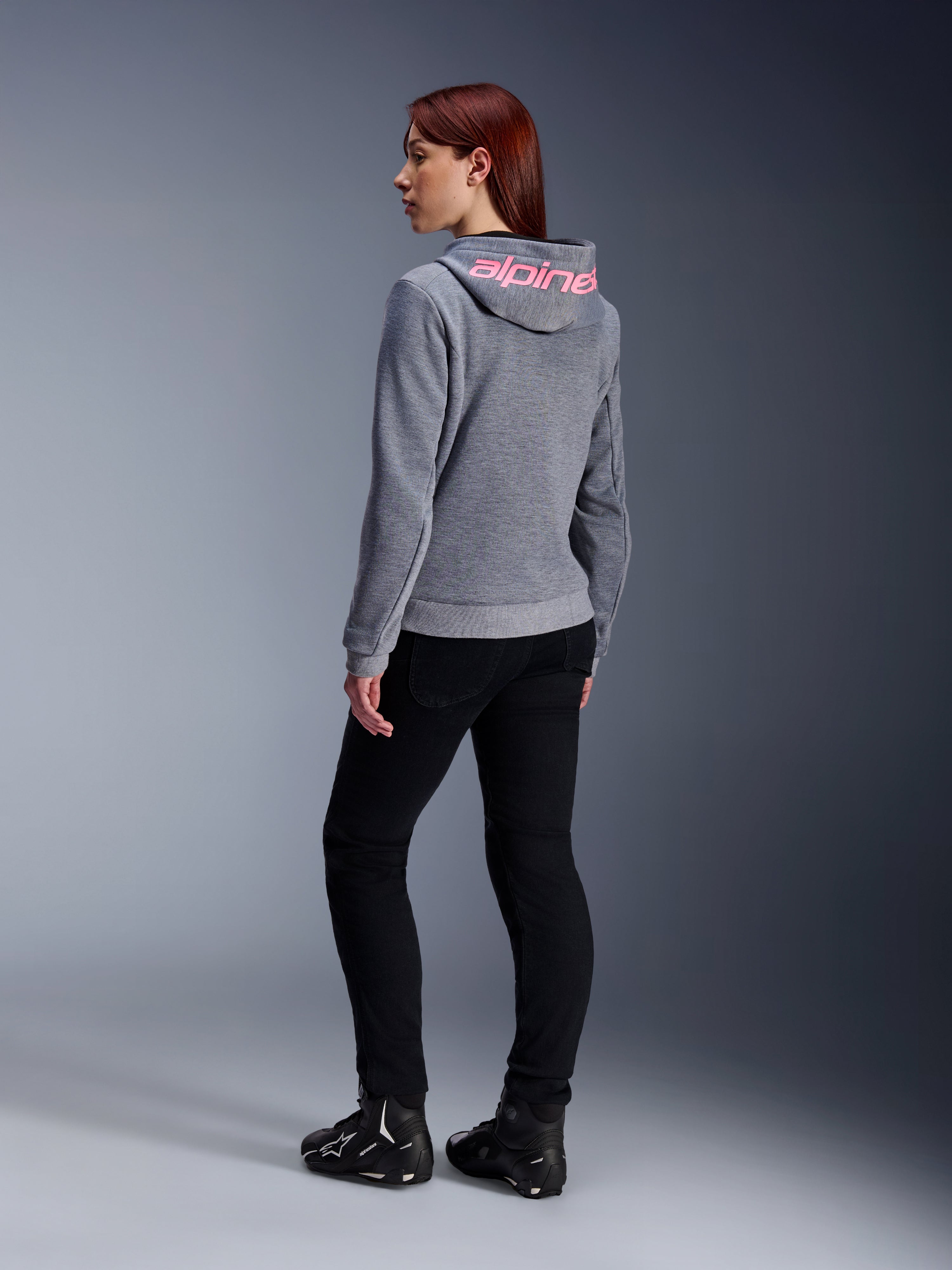 Alpinestars Stella Chrome V2 Sport Hoodie, Damen-Motorrad-Hoodie, Grau meliert mit pinkem Fluo-Branding auf der Kapuze, Rückansicht, kombiniert mit schwarzer Hose und Motorradschuhen, lässiger urbaner Street-Style mit Motorradschutz