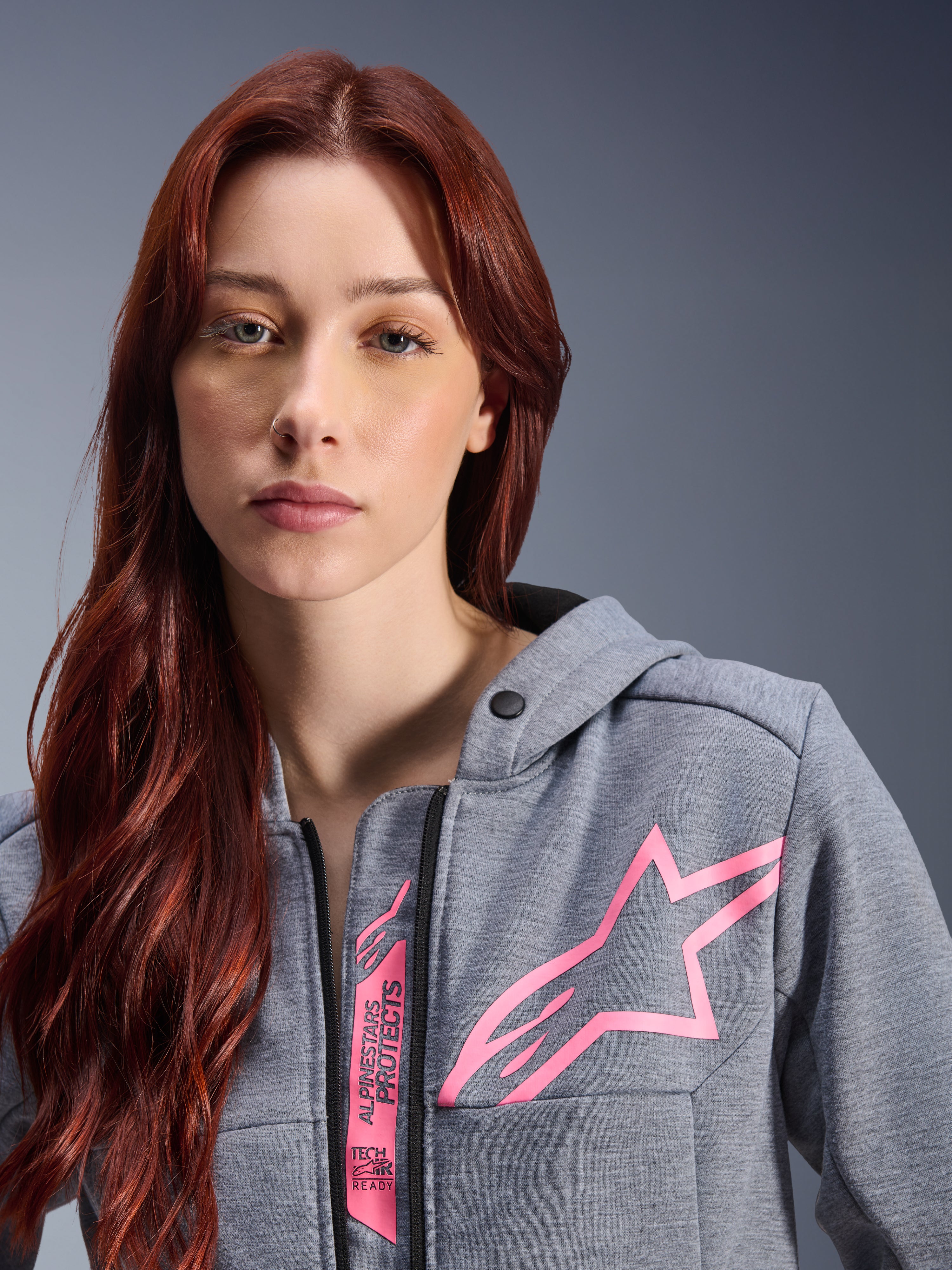 Frau trägt Alpinestars Stella Chrome V2 Sport Hoodie, Grau Meliert Pink Fluo, Nahaufnahme mit dem pinkfarbenen Alpinestars Stern-Logo und Tech-Air Ready Branding, urbane Motorradbekleidung.