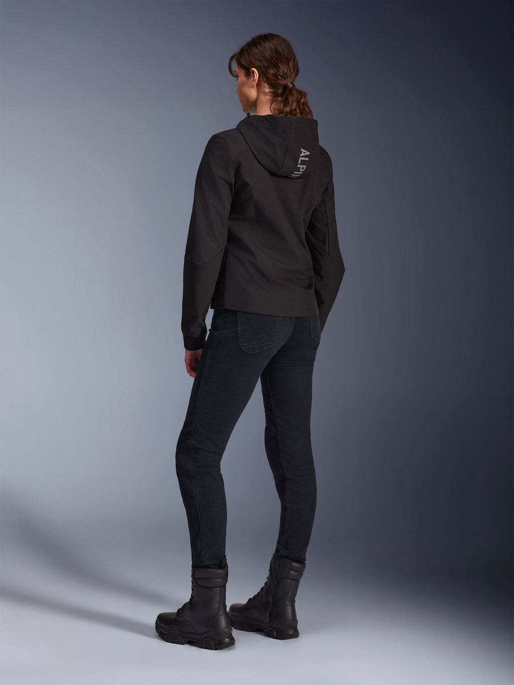 Alpinestars Stella Radium Tech Hoodie, schwarzer Urban-Riding-Hoodie für Damen, Rückansicht mit „ALPIN“-Branding auf der Kapuze, kombiniert mit schwarzer technischer Motorradhose und schwarzen Stiefeln, leichte winddichte Tech-Shell.