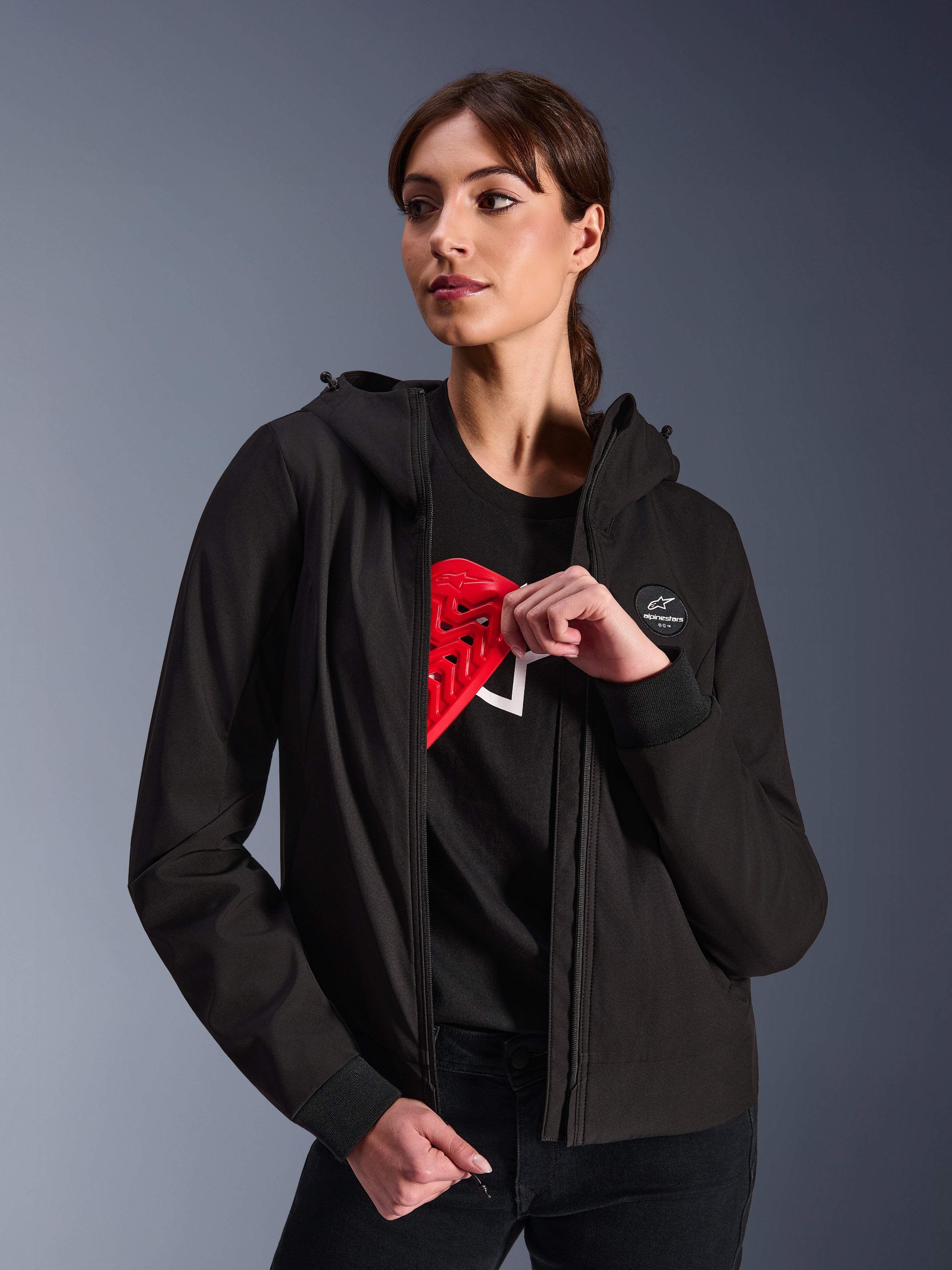Alpinestars Stella Radium Tech Hoodie für Damen, schwarzer Urban-Riding-Hoodie, weibliches Model demonstriert rote Nucleon Flex Plus Protektoren im Inneren, leichtes 3-lagiges winddichtes Textil-Außenmaterial mit gerippten Bündchen und Logo-Patch auf der Brust, schützende Motorradbekleidung
