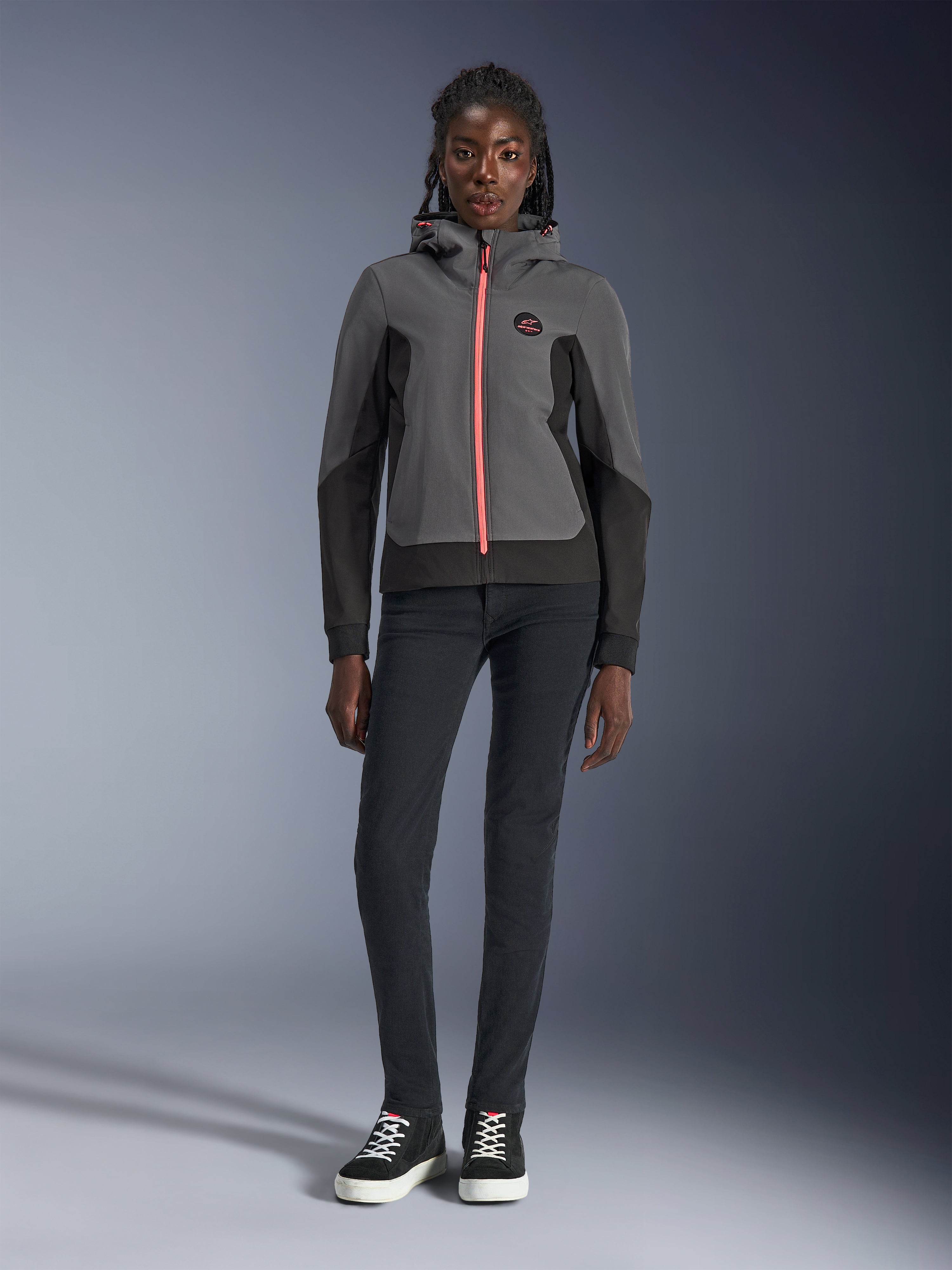 Female model wearing the Alpinestars Stella Radium Tech Hoodie, Deep Gray and Black with a bright orange zipper accent, urban motorcycle riding hoodie, paired with black slim-fit pants and sneakers, casual athletic performance gear for women.