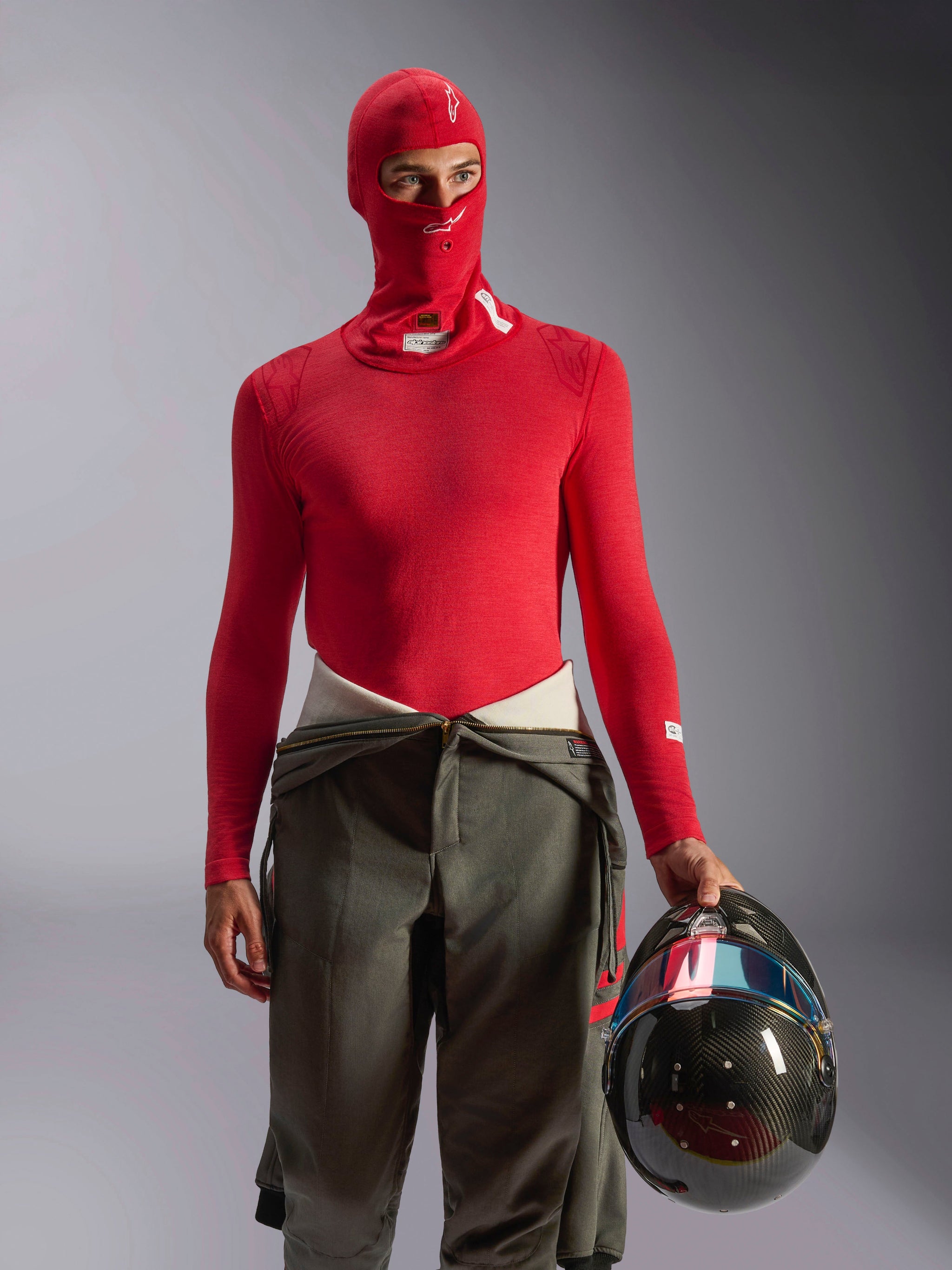 Alpinestars ZX Evo V3 Balaclava in Red, auto racing base layer worn by driver with matching red long-sleeve top and olive green racing pants, holding a carbon fiber helmet, professional flame-resistant motorsport gear