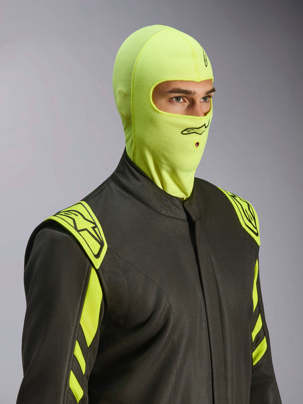 Male model wearing Alpinestars ZX Evo V3 Balaclava in Yellow Fluo, flame-resistant racing head sock, paired with a dark grey auto racing suit with neon yellow accents, featuring a black Alpinestars logo and mouth opening for hydration tube, professional motorsport protective gear.