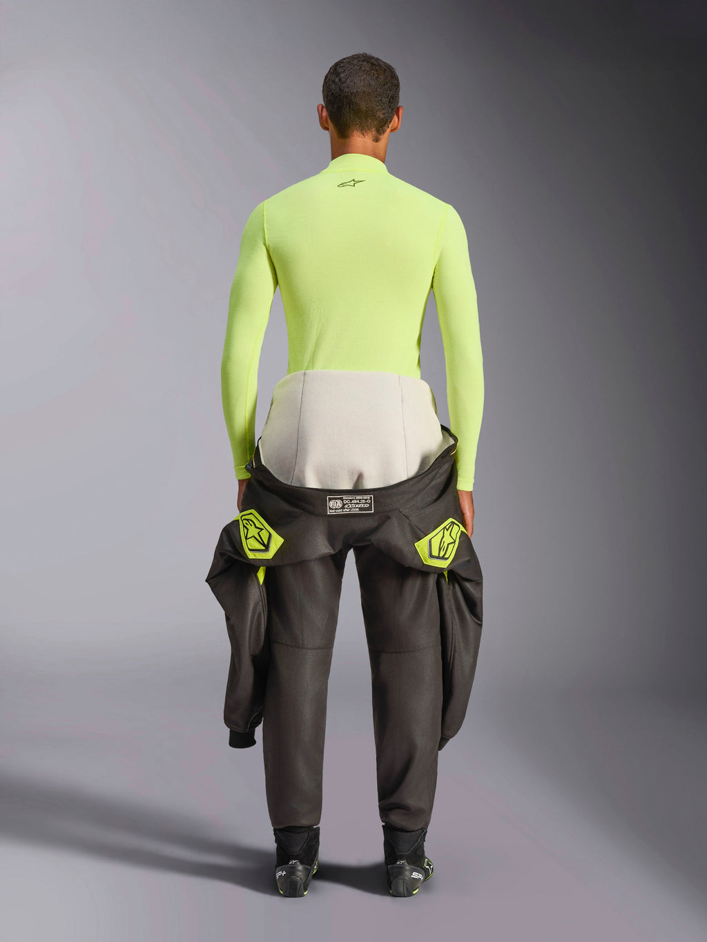 Alpinestars ZX Evo V3 Top, Langarm-Base-Layer, Yellow Fluo, Rückansicht eines Fahrers mit um die Taille gebundenem Rennanzug, leuchtend neongelbes, flammhemmendes Oberteil mit kleinem schwarzen Logo am Nacken und athletischer Passform