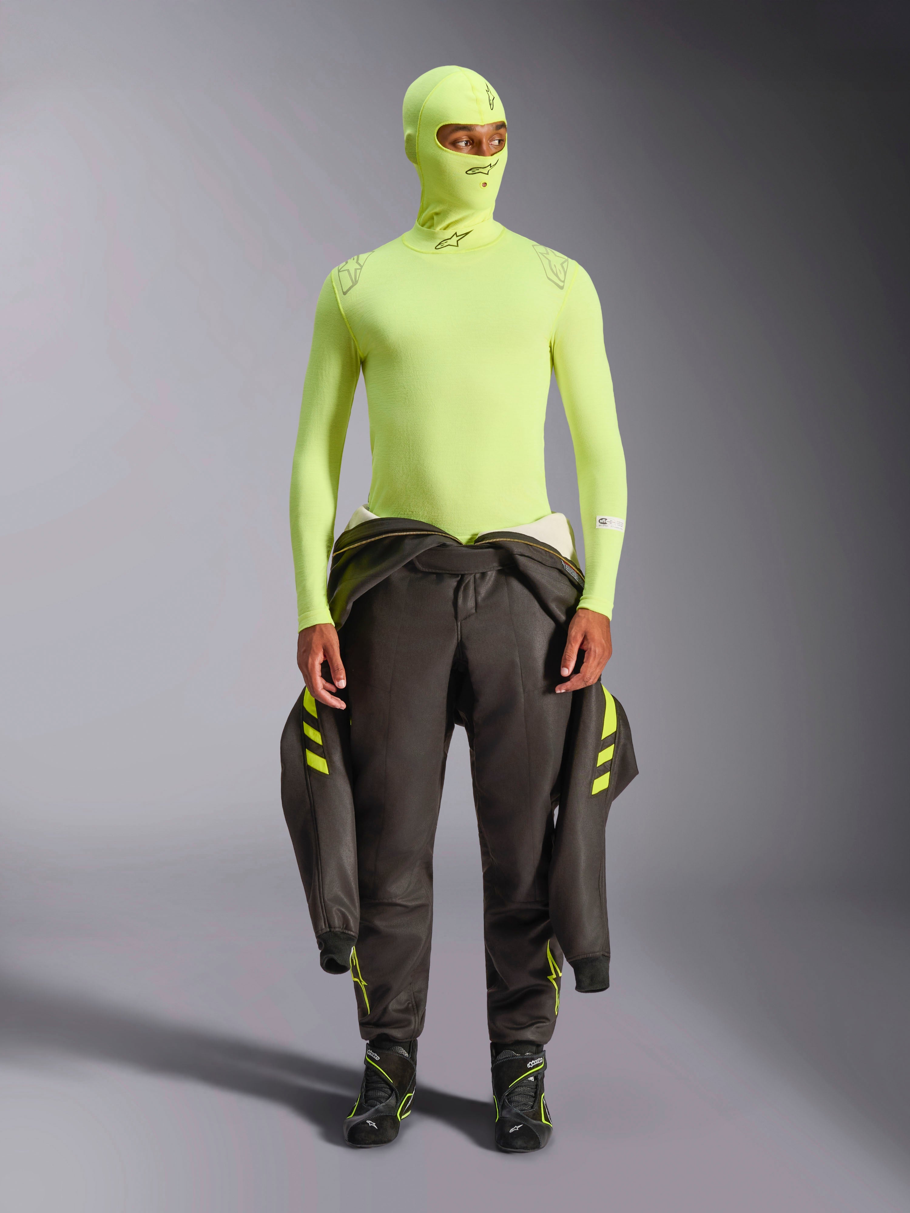 Alpinestars ZX Evo V3 Langarm-Oberteil, Yellow Fluo, getragen mit passender Sturmhaube und einem um die Taille gebundenen dunklen Rennanzug, leichte flammhemmende Basisschicht für den Autorennsport, mit Alpinestars-Logos an Kragen und Schultern