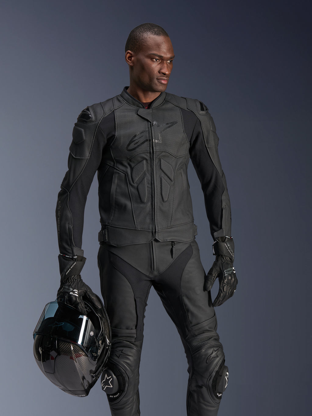 Alpinestars Dusk Airflow Lederjacke, Schwarz, getragen von einem männlichen Fahrer mit passender Lederhose und Handschuhen, hält einen schwarzen Helm, mit perforierten Lederbahnen und integrierten Protektoren für sportliches Motorradfahren