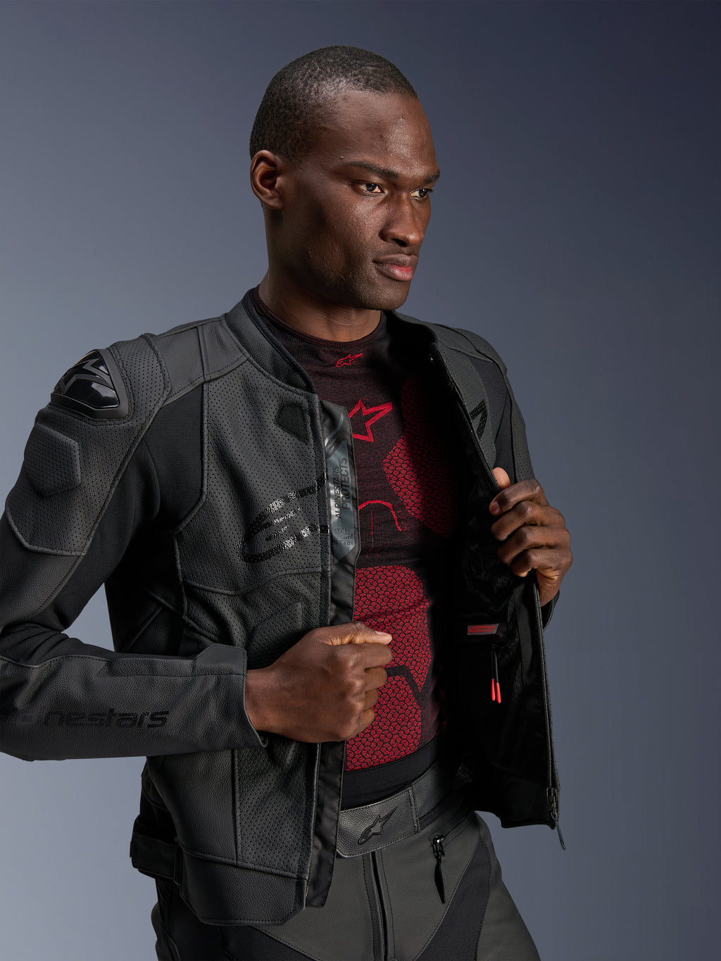 Alpinestars Dusk Airflow Lederjacke, Black Black, getragen von einem männlichen Modell über einer rot-schwarzen Basisschicht, mit perforierten Ledereinsätzen, externen DFS-Schulterschleifern und einer sportlichen Performance-Passform.