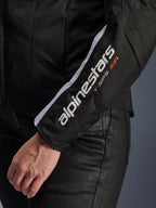 Alpinestars Stella T-SPS Air V2 Jacke, Motorradjacke für Damen, Schwarz und Weiß, Nahaufnahme des Ärmels mit weißem Alpinestars-Logo und T-SPS AIR-Schriftzug, Mesh- und Textilkonstruktion für Luftzirkulation, getragen mit schwarzer Motorradhose