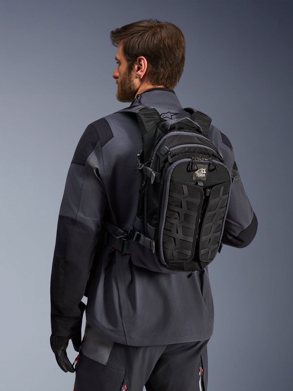 Alpinestars AT-15 Rucksack, Adventure-Motorradrucksack, Schwarz und Dunkelgrau, getragen von einem Fahrer in einer grauen Jacke, mit technischem Design, MOLLE-Gurtband und „TERRA“-Branding, ergonomische Offroad-Ausrüstung