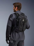 Alpinestars AT-15 Rucksack, Adventure-Motorradrucksack, Schwarz und Dunkelgrau, getragen von einem Fahrer in einer grauen Jacke, mit technischem Design, MOLLE-Gurtband und „TERRA“-Branding, ergonomische Offroad-Ausrüstung