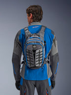 Alpinestars AT-15 Back Pack, adventure motorcycle backpack, Dark Gray and Deep Gray, worn by a rider over a blue and gray jacket, featuring All Terra branding, ergonomic structural design, adjustable straps, and blue zipper accents.