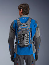Alpinestars AT-15 Rucksack, Adventure-Motorradrucksack, Dunkelgrau und Tiefgrau, getragen von einem Fahrer über einer blau-grauen Jacke, mit All-Terra-Branding, ergonomischem strukturellem Design, verstellbaren Riemen und blauen Reißverschluss-Akzenten.