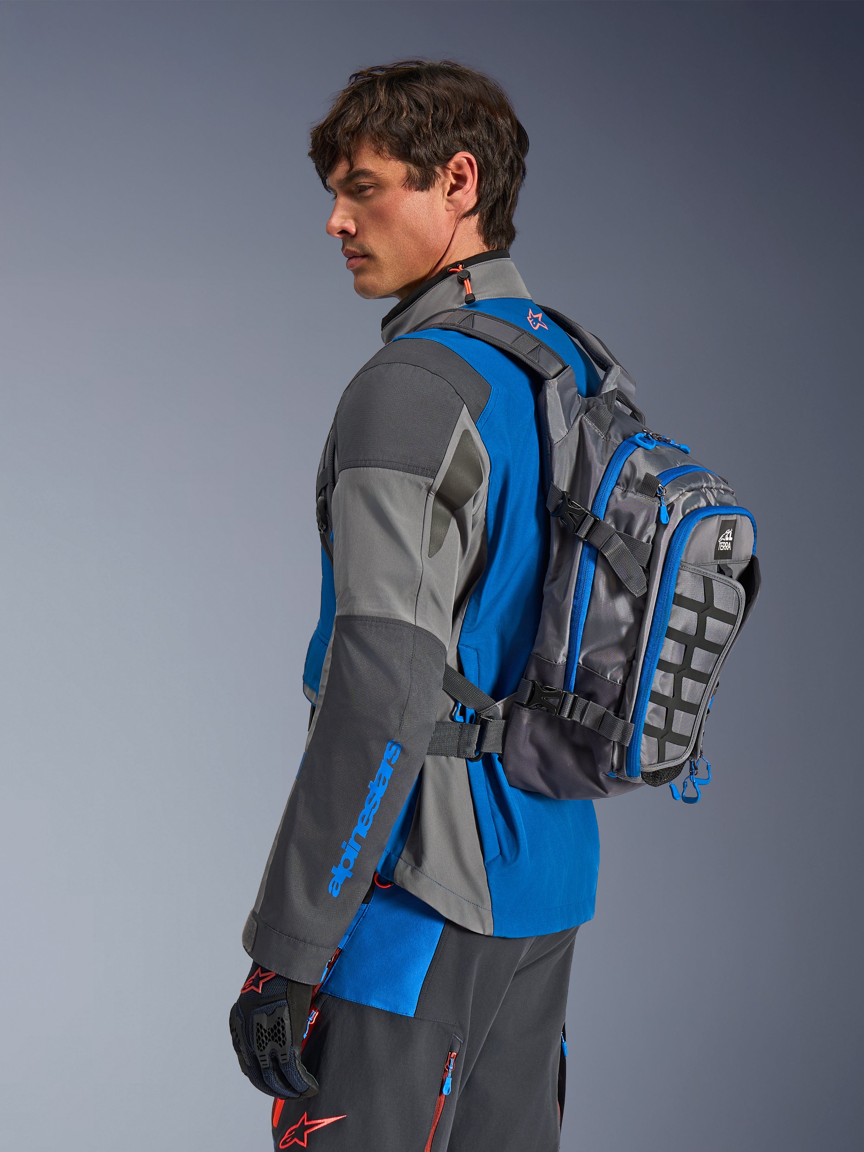 Alpinestars AT-15 Back Pack, Motorcycle Adventure Backpack, Dark Gray with Blue accents, worn by a rider in coordinated gray and blue technical riding gear, featuring MOLLE panel and rugged construction for off-road adventure.