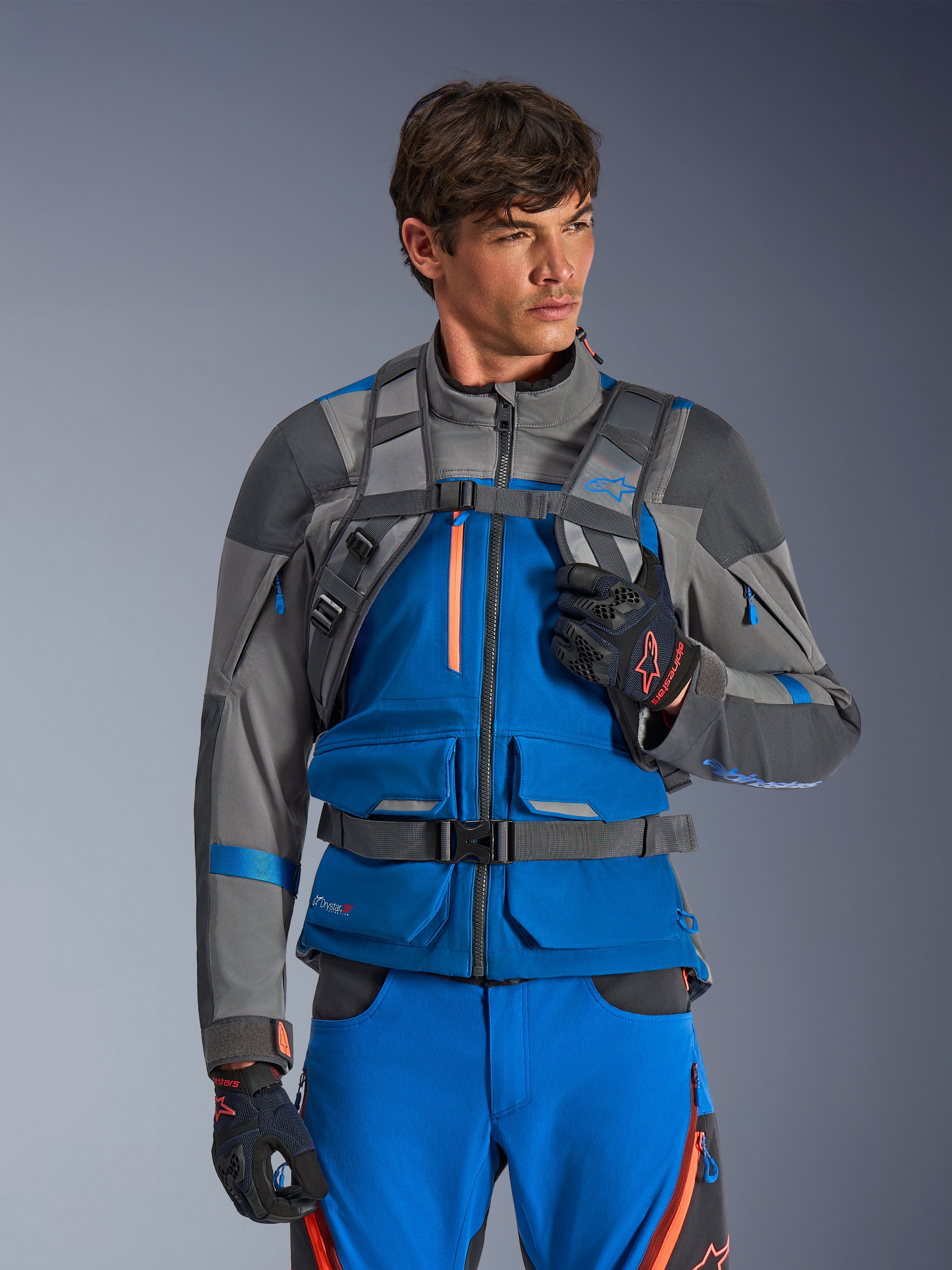 Alpinestars AT-15 Back Pack, Motorcycle Backpack, Dark Gray, worn by a rider in a blue and gray adventure jacket and pants, featuring technical gray shoulder straps with magnetic chest buckle and waist strap, rugged design for off-road riding