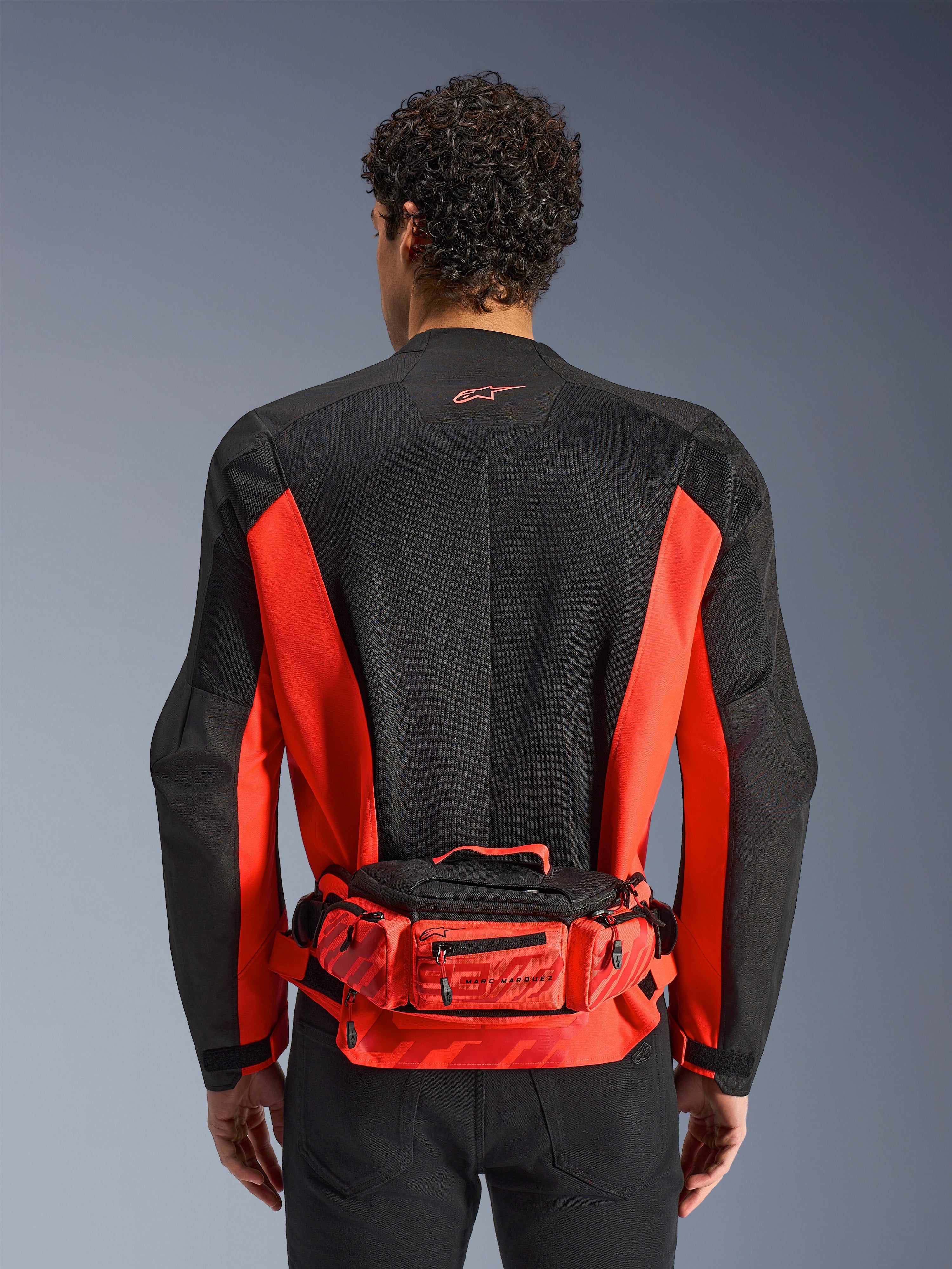 Alpinestars MM93 Hüfttasche, Rot und Schwarz, getragen über einer schwarz-roten Motorradjacke, mit Marc Marquez 93 Branding, verstellbarem Gurt und Reißverschlussfächern, funktionales, vom Rennsport inspiriertes Accessoire