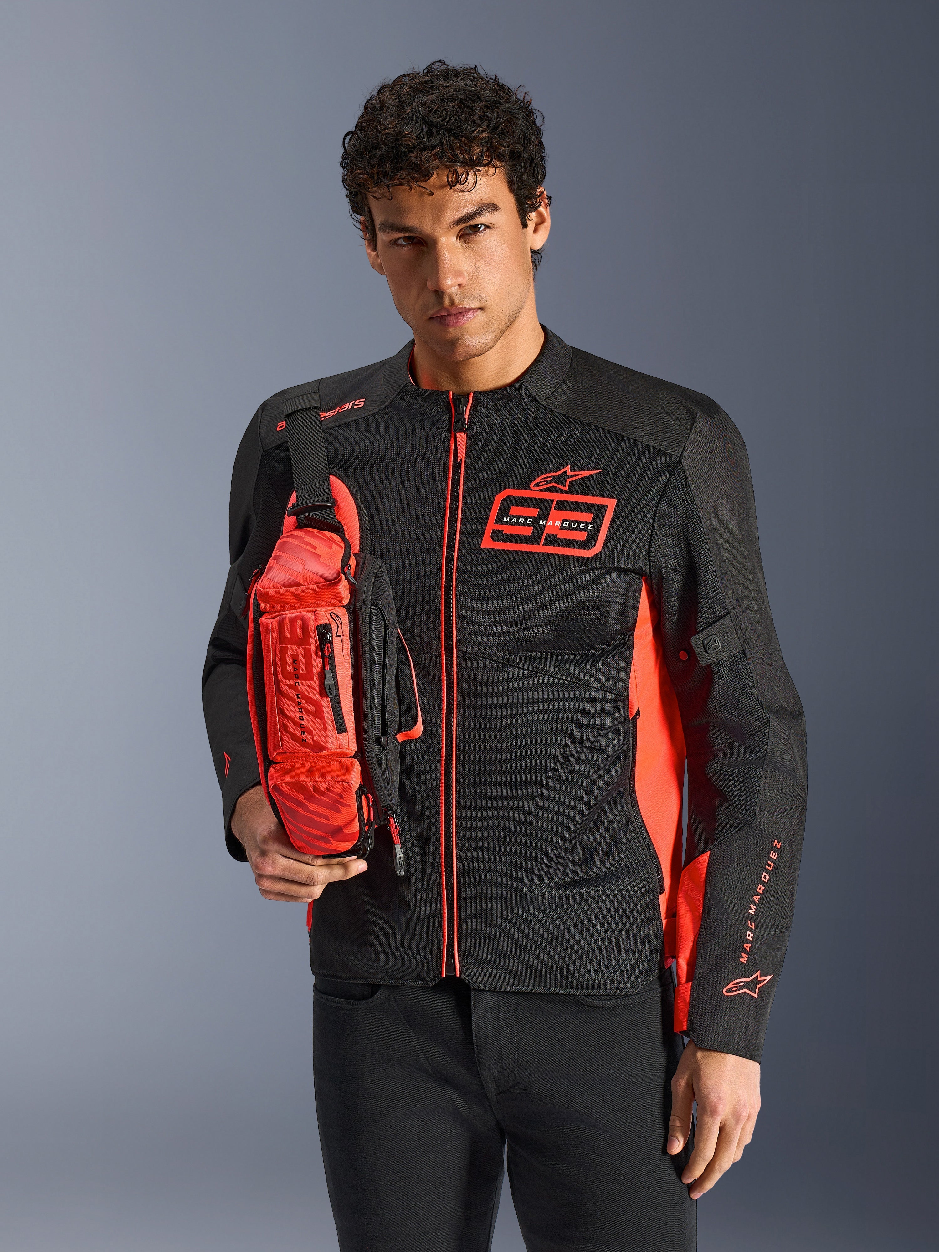 Mann mit Alpinestars MM93 Waist Bag, Rot Fluo und Schwarz, als Crossbody-Tasche über einer passenden schwarz-roten Motorradjacke getragen, mit Marc Marquez 93-Logo, sportliches Racing-Design