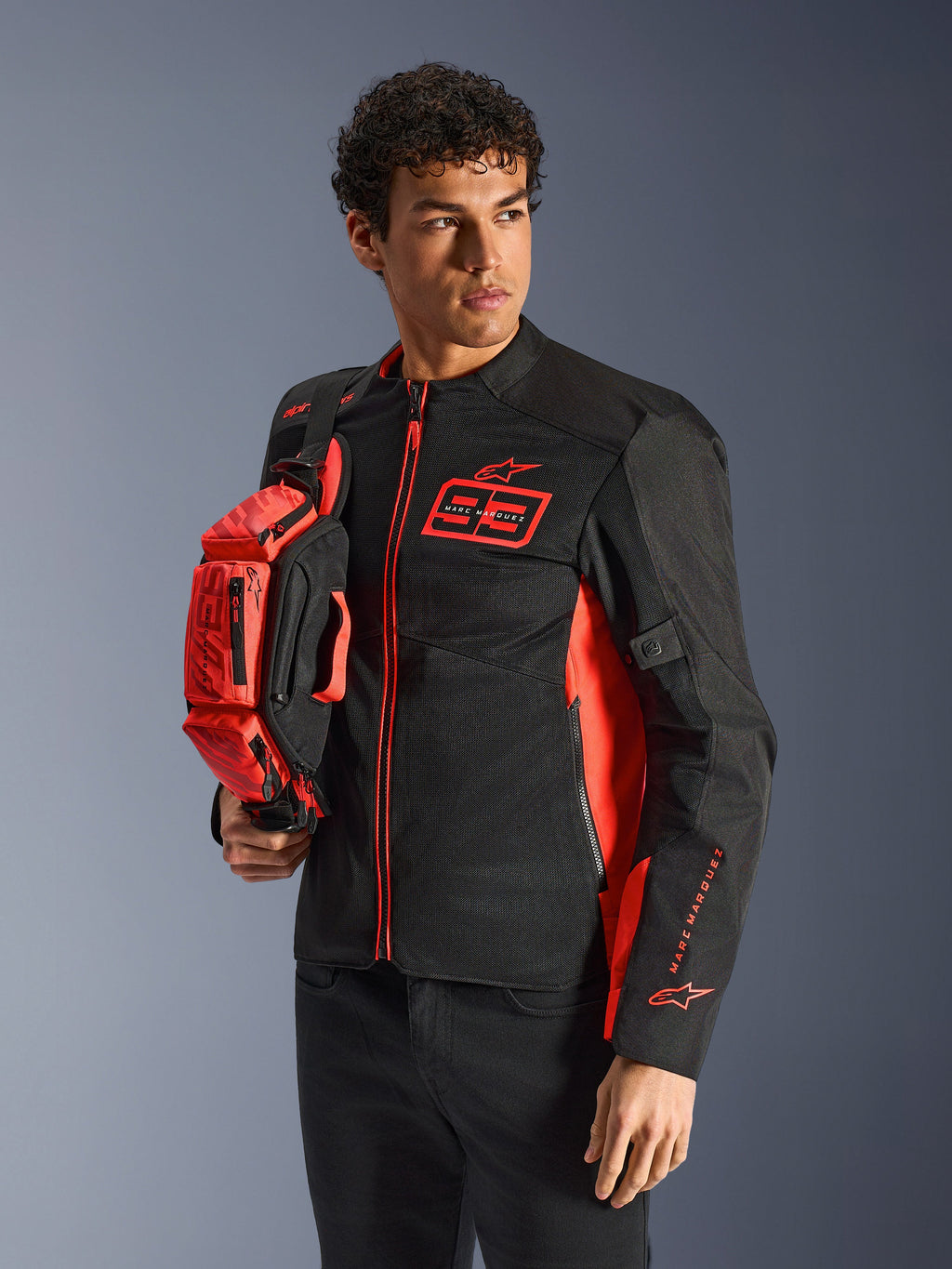 Alpinestars MM93 Hüfttasche, Red Fluo und Schwarz, gehalten von einem Model in einer passenden schwarz-roten Motorradjacke, mit mehreren Reißverschlussfächern und Marc Marquez Branding, praktische Ausrüstung für sportliches Fahren