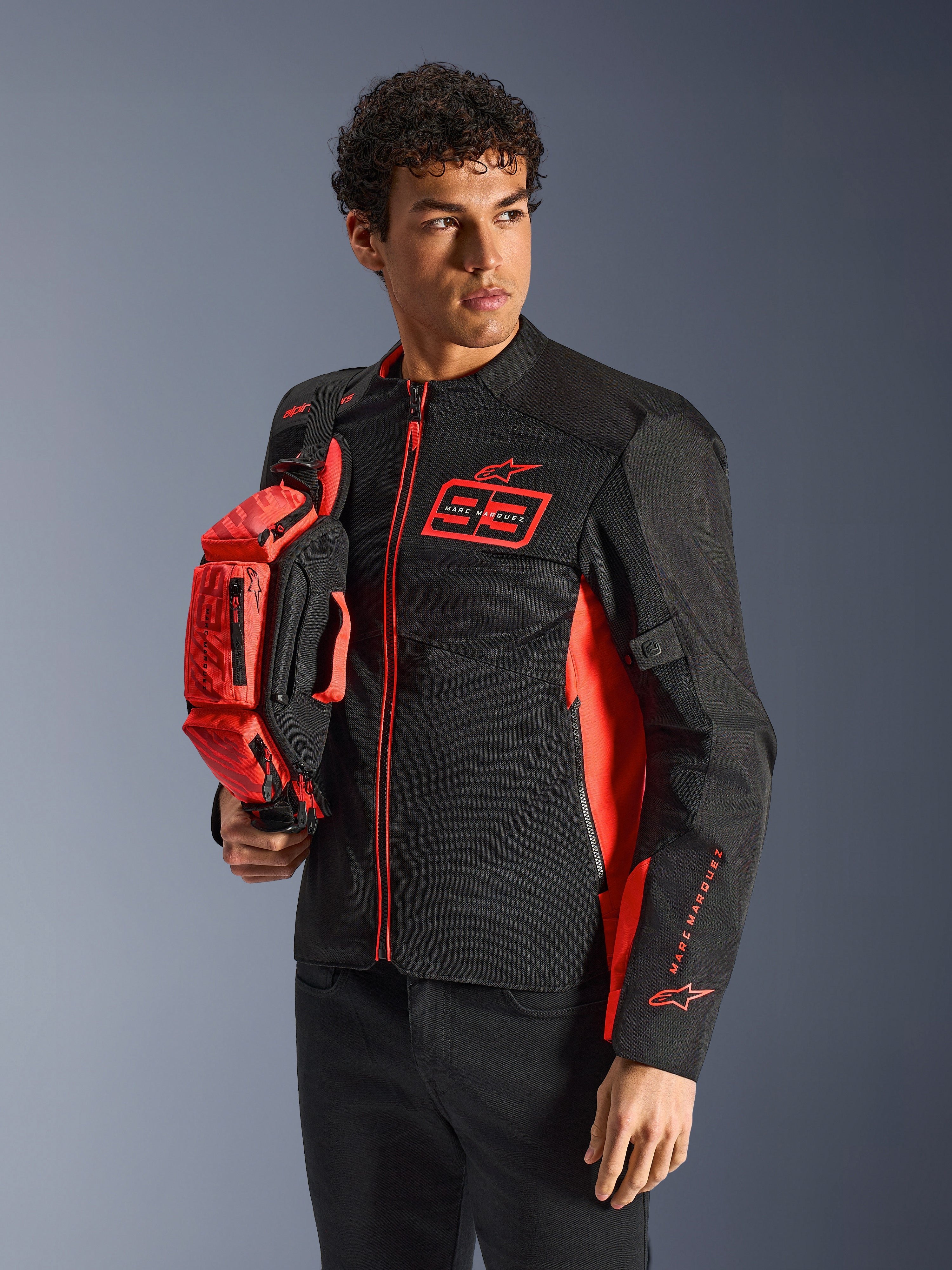 Alpinestars MM93 Hüfttasche, Red Fluo und Schwarz, gehalten von einem Model in einer passenden schwarz-roten Motorradjacke, mit mehreren Reißverschlussfächern und Marc Marquez Branding, praktische Ausrüstung für sportliches Fahren