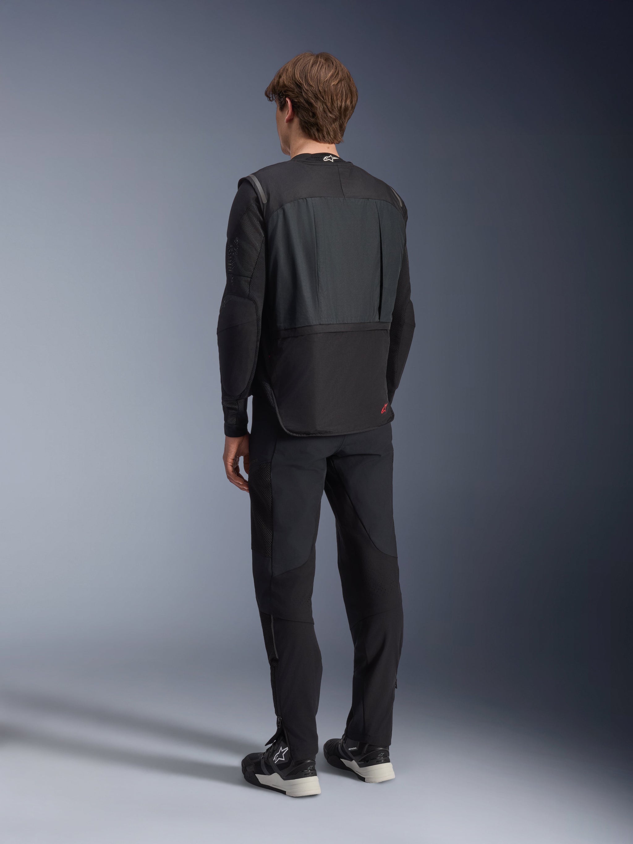 Alpinestars Tech-Air 3 V2 System, Black autonomous motorcycle airbag vest, rear view worn by a model with black riding pants, features a lightweight and slim profile, red logo on lower back, advanced rider safety gear for touring and commuting