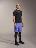 Male model wearing Alpinestars A-Aria Switch Short Sleeve Jersey in Black with white chest branding, paired with light blue mountain bike shorts, black knee guards, and black cycling shoes, studio full-body shot for performance MTB gear