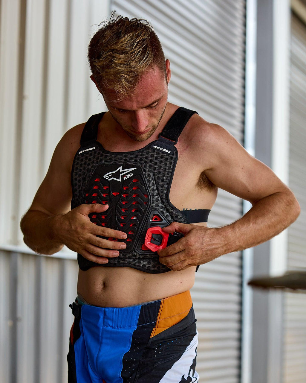 Mann beim Anlegen des Alpinestars A-5 Plasma Brustprotektors, Schwarz mit roten Akzenten und weißem Logo, Waben-Netzstruktur, kombiniert mit blau-orangen Motocross-Hosen, Vorbereitung auf ein Offroad-Rennen