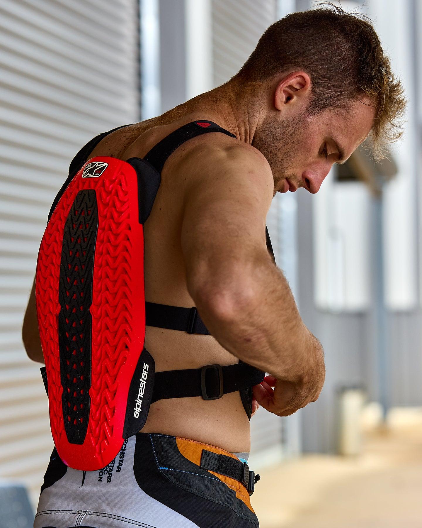 Alpinestars A-5 Plasma Brustprotektor, Rot und Schwarz, mit strukturiertem Rückenprotektor und Nucleon PLASMA-Technologie, getragen von einem Mann beim Anpassen der schwarzen Gurte, Seitenansicht zeigt die Schutzschale und das flexible Design für Offroad-Fahrten