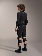 Modell trägt Alpinestars A-Impact Plasma Pro Knie-Schienbein-Protektoren in der Rückansicht, Schwarz und Weiß, zeigt den Dreifach-Riemenverschluss und belüftete Mesh-Einsätze, kombiniert mit einer kompletten schwarzen Mountainbike-Schutzausrüstung inklusive langärmeligem Baselayer und gepolsterten Shorts