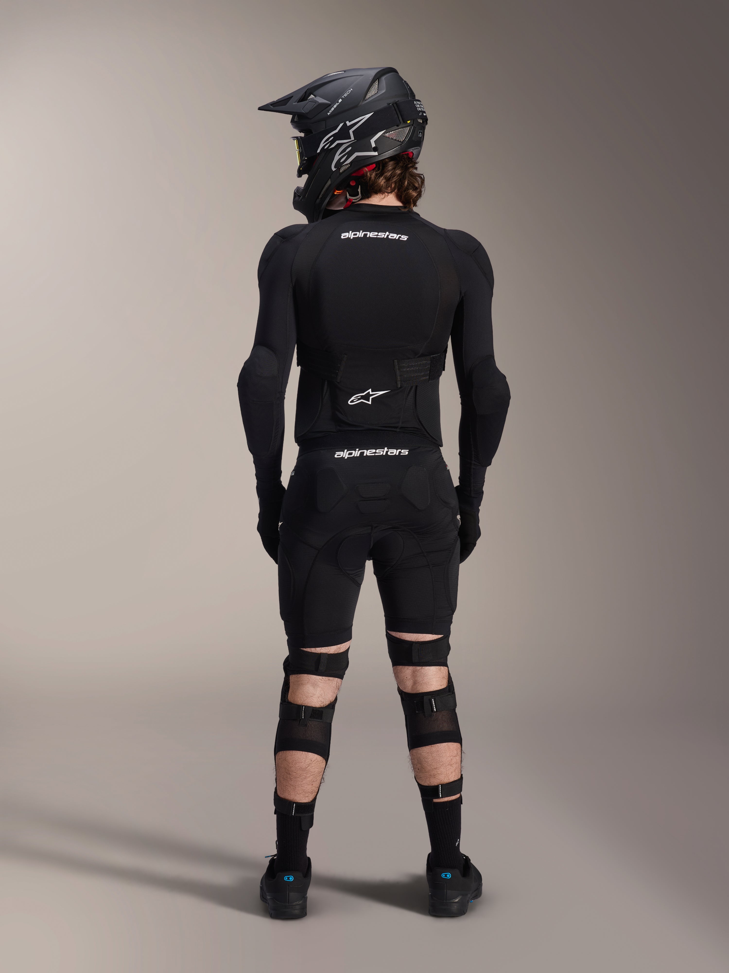 Alpinestars A-Impact Plasma Pro Knie-Schienbeinschützer, Schwarz, Rückansicht eines Fahrers in kompletter Mountainbike-Schutzausrüstung inklusive Fullface-Helm und Body Armor, mit Dreifach-Riemen-Verschlusssystem und belüfteten Mesh-Einsätzen für sicheren Sitz und Luftzirkulation
