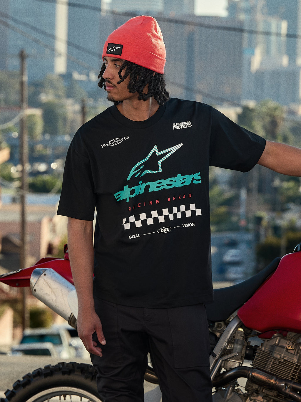 Alpinestars Aplomb Oversized CSF Tee, Kurzarm-T-Shirt, Schwarz, mit großem blaugrünem und weißem Astars-Logo mit Farbverlauf und Zielflaggen-Grafik, getragen mit einer korallenfarbenen Beanie, stehend neben einem roten Motorrad, lässige Lifestyle-Streetwear