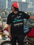 Alpinestars Aplomb Oversized CSF Tee, Kurzarm-T-Shirt, Schwarz, mit großem blaugrünem und weißem Astars-Logo mit Farbverlauf und Zielflaggen-Grafik, getragen mit einer korallenfarbenen Beanie, stehend neben einem roten Motorrad, lässige Lifestyle-Streetwear