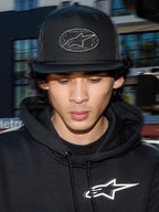 Alpinestars Contender Trucker Hat, schwarze Lifestyle-Cap mit Poly-Mesh-Rückseite, strukturierte Krone mit einem bestickten Astars-Logo-Patch, getragen von einem Mann mit einem schwarzen Alpinestars-Hoodie, lässige, vom Motorsport inspirierte Streetwear.