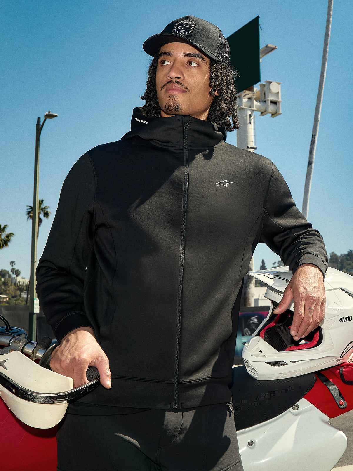 Alpinestars Cypher Hoodie, schwarzes Lifestyle-Fleece mit Reißverschluss, hochgeschlossene Kapuze mit Branding, kleines weißes Astars-Logo auf der Brust, getragen mit einer schwarzen Kappe von einem Fahrer, der einen weißen Motocross-Helm hält, lässige Motorsport-Streetwear