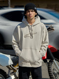 Alpinestars Emergent Oversized Hoodie in Off White, Oversized-Fleece-Hoodie mit 3D-besticktem Astars-Logo, Kängurutasche und Kapuze mit Kordelzug, getragen von einem Mann mit schwarzer Beanie neben einem Motorrad, lässige Lifestyle-Motorsport-Streetwear