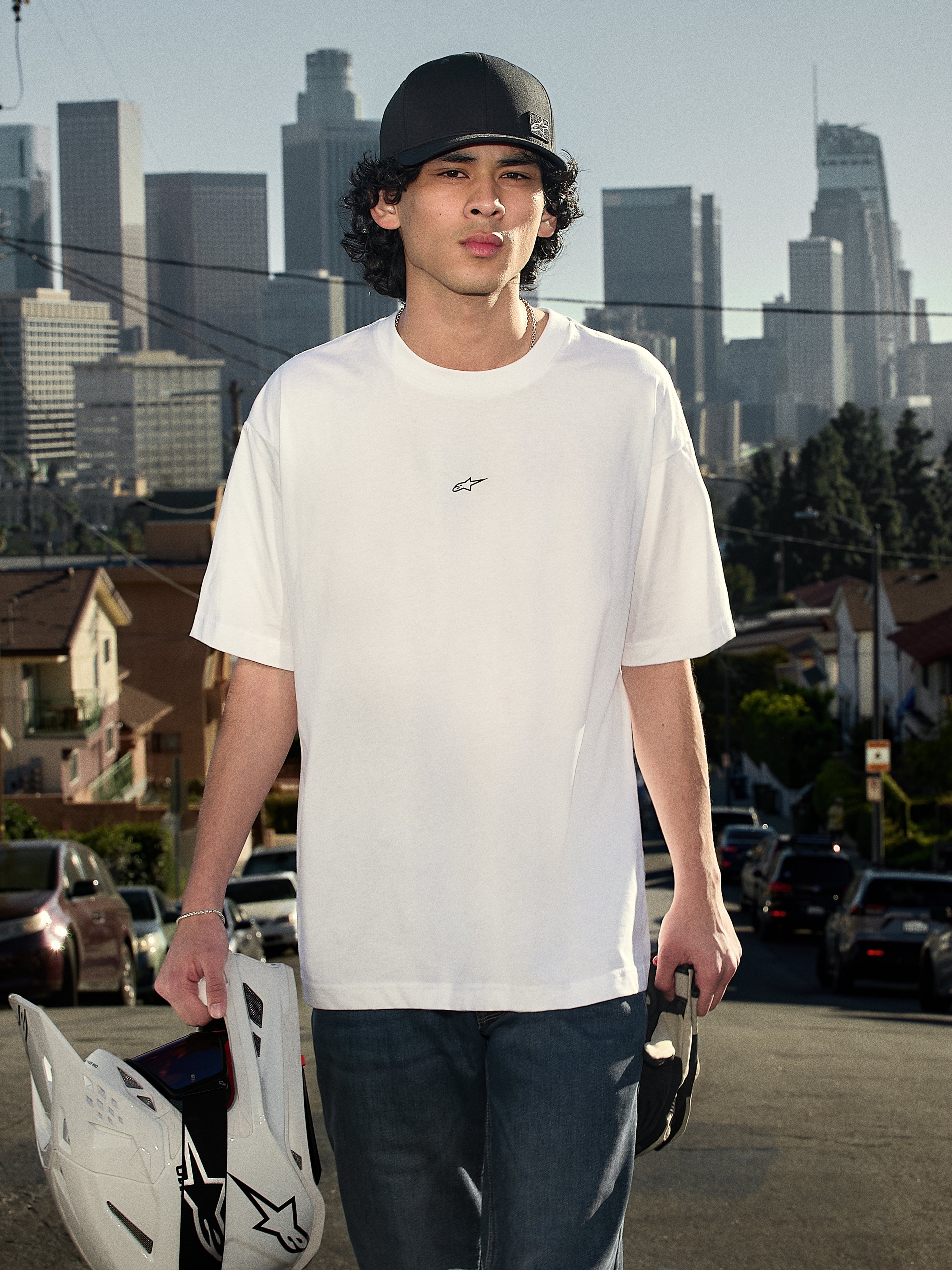 Alpinestars Focused Oversized CSF Tee, kurzärmeliges T-Shirt, Weiß, mit einem kleinen schwarzen Astars-Logo auf der Brust, Lifestyle-Aufnahme einer Person, die das T-Shirt trägt und einen Motocross-Helm hält, Skyline im Hintergrund, lässige Motorsport-Streetwear.
