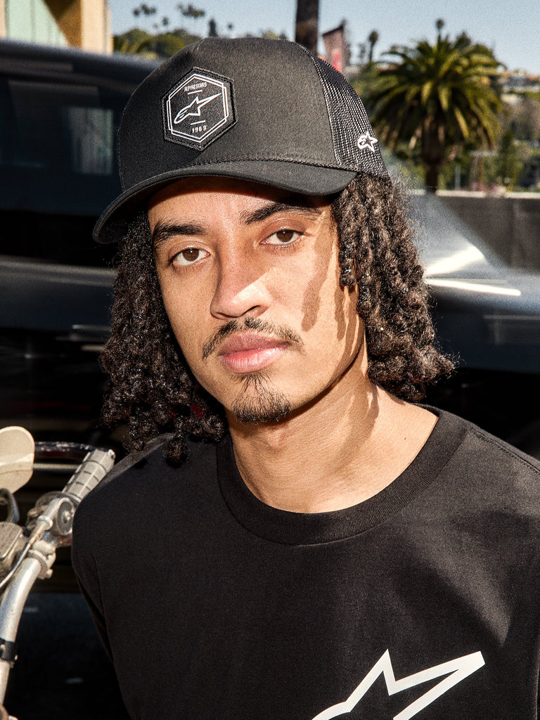 Alpinestars Hexeon Trucker Hat, Black, lifestyle shot of a man wearing the curved bill trucker cap with hexagonal Astars logo patch and mesh back, paired with a black logo tee, casual motorsport-inspired streetwear