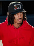 Mann mit Alpinestars Intuitive Snapback-Kappe in Schwarz, mit strukturierter Krone und 'Racing Ahead' Logo-Stickerei-Patch, kombiniert mit einem leuchtend roten Hoodie, lässige Lifestyle-Streetwear