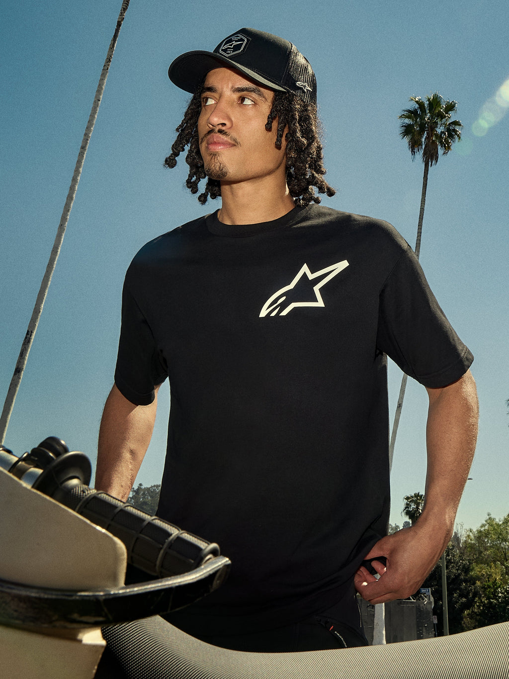Alpinestars One Oversized CSF Tee, Kurzarm-T-Shirt, Schwarz mit weißem Astars-Logo, getragen von einem Mann mit schwarzer Trucker-Cap, Outdoor-Lifestyle-Setting mit Palmen und einem Motorrad, lässige Motorsport-Streetwear