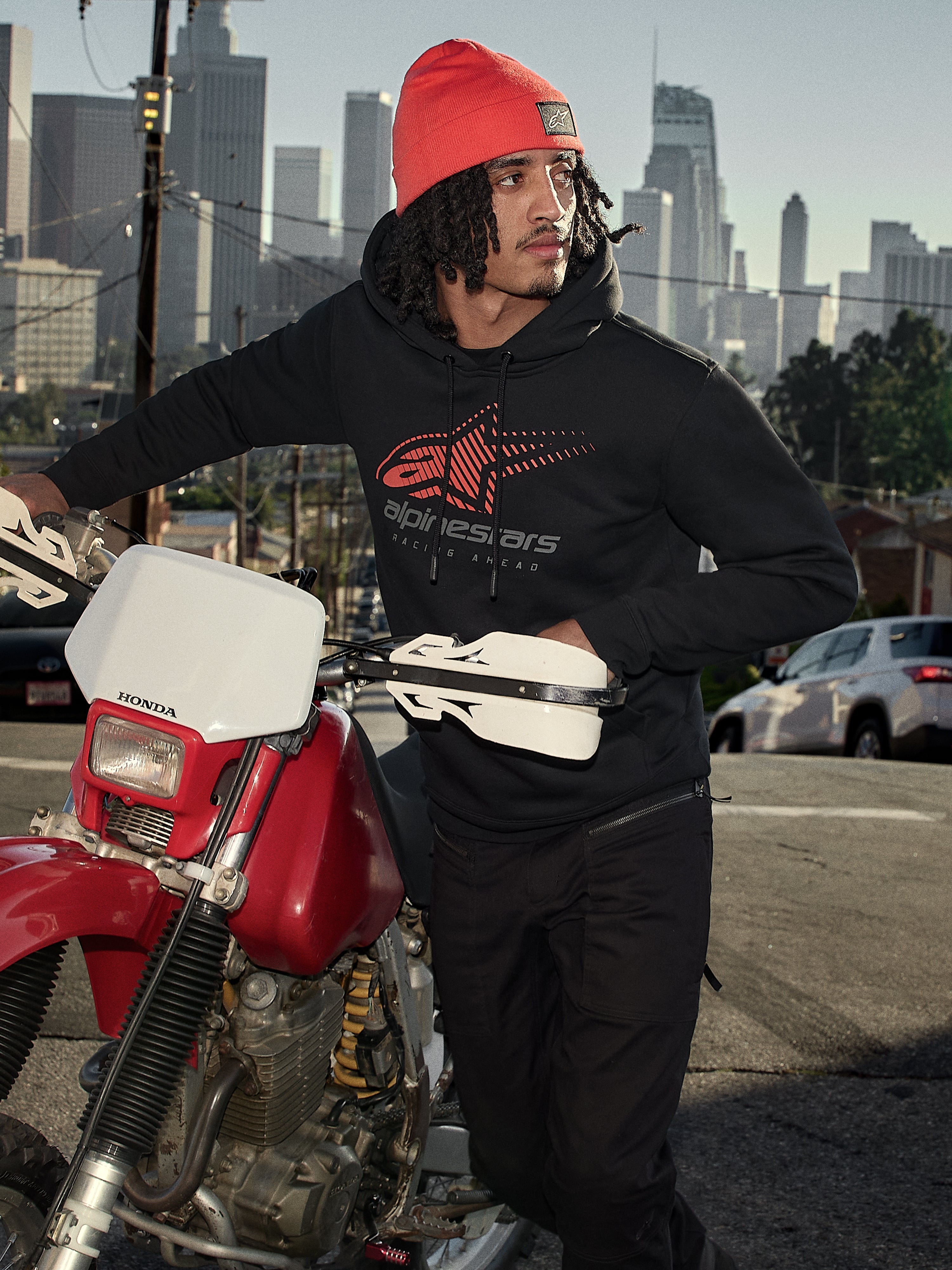 Alpinestars Pulse Hoodie, Schwarz, Kapuzenpullover aus Fleece mit rot-grauem Astars Combo Logo, getragen von einem Mann mit orangefarbener Beanie neben einem roten Honda-Motorrad in einer städtischen Umgebung, lässige Motorsport-Streetwear